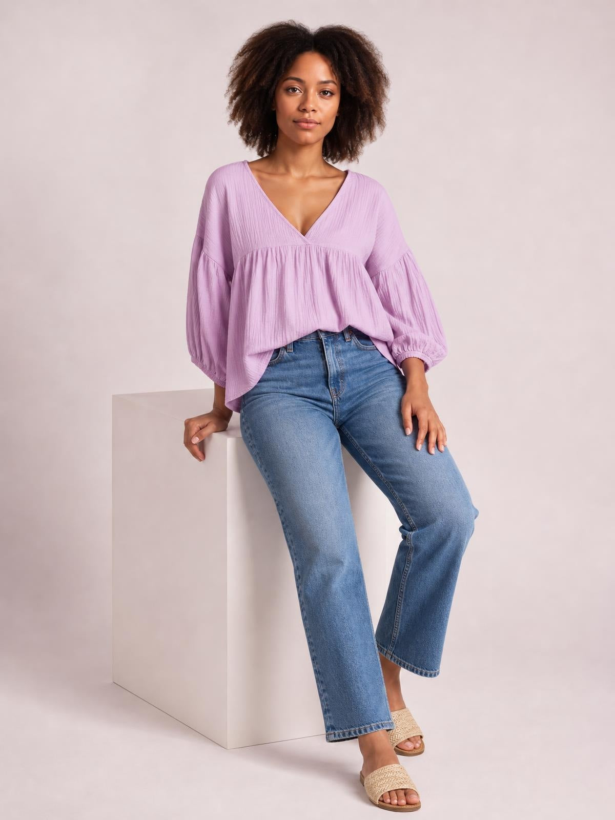 Model wears a lavender dropped-shoulder V-neck blouse with long gathered sleeves and a loose, flowing silhouette, seated casually with one hand resting beside her.