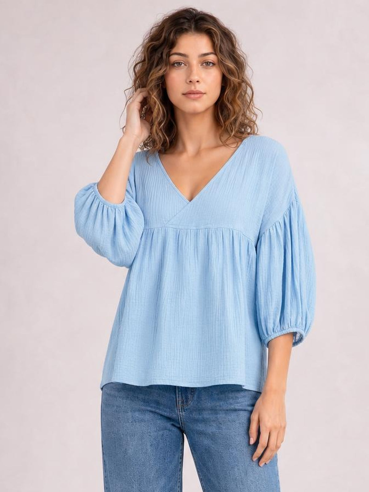 A model poses in a light blue solid V-neck blouse with dropped shoulders, long gathered sleeves, a tiered relaxed silhouette, and a long hem.