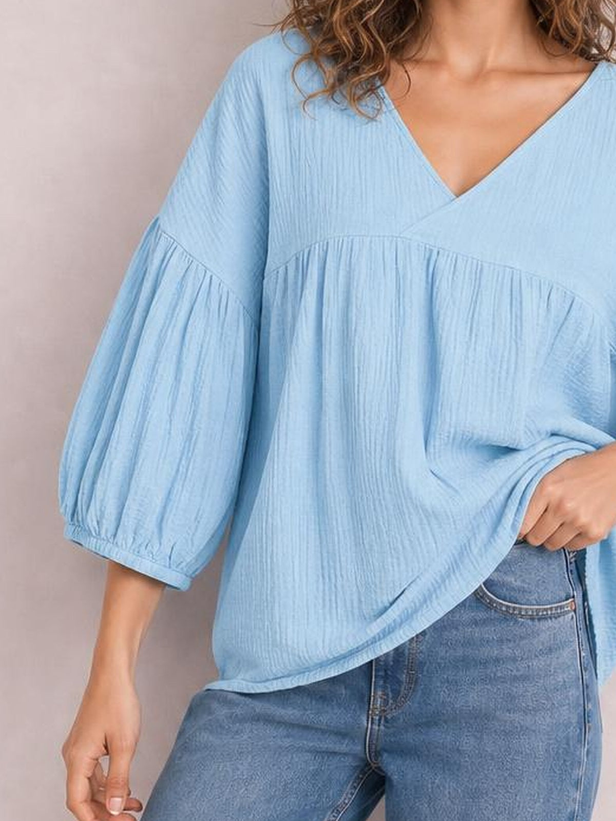 Model wearing a solid light blue dropped shoulder V-neck blouse with long voluminous sleeves, gathered seam detailing, and a relaxed long silhouette.