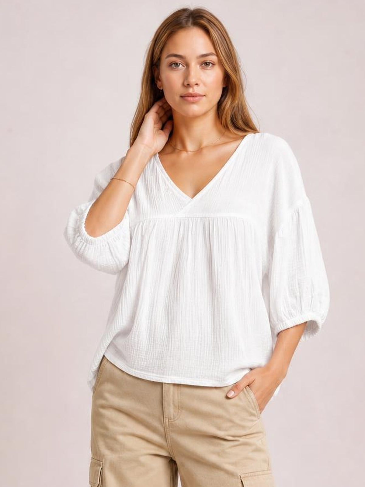 Model wearing a solid white dropped shoulder V-neck blouse with long gathered sleeves, elastic cuffs, a relaxed long silhouette, and textured fabric.
