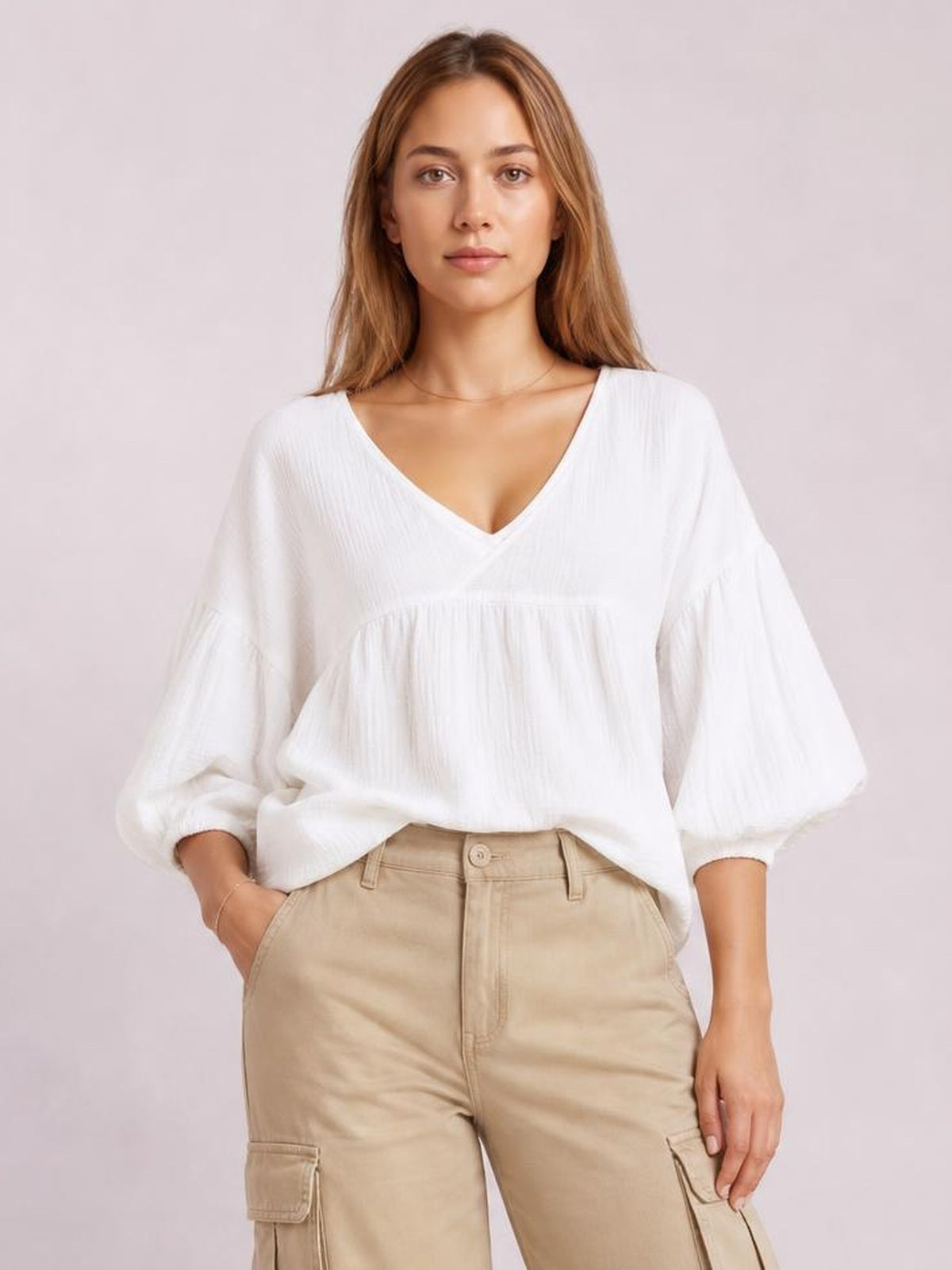 Model wearing a solid white dropped shoulder V-neck blouse with long sleeves, gathered panel construction, and a relaxed, long silhouette.