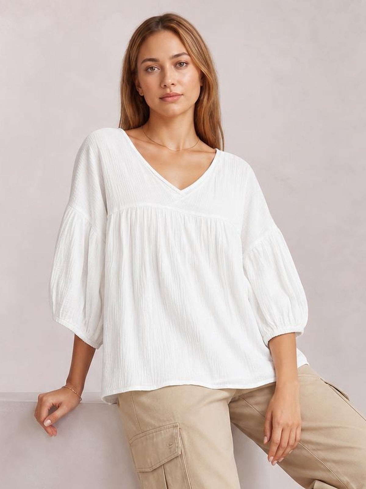 A model sits wearing a solid white dropped-shoulder V-neck blouse with long sleeves, a relaxed flowing silhouette, and a gathered seam across the chest.