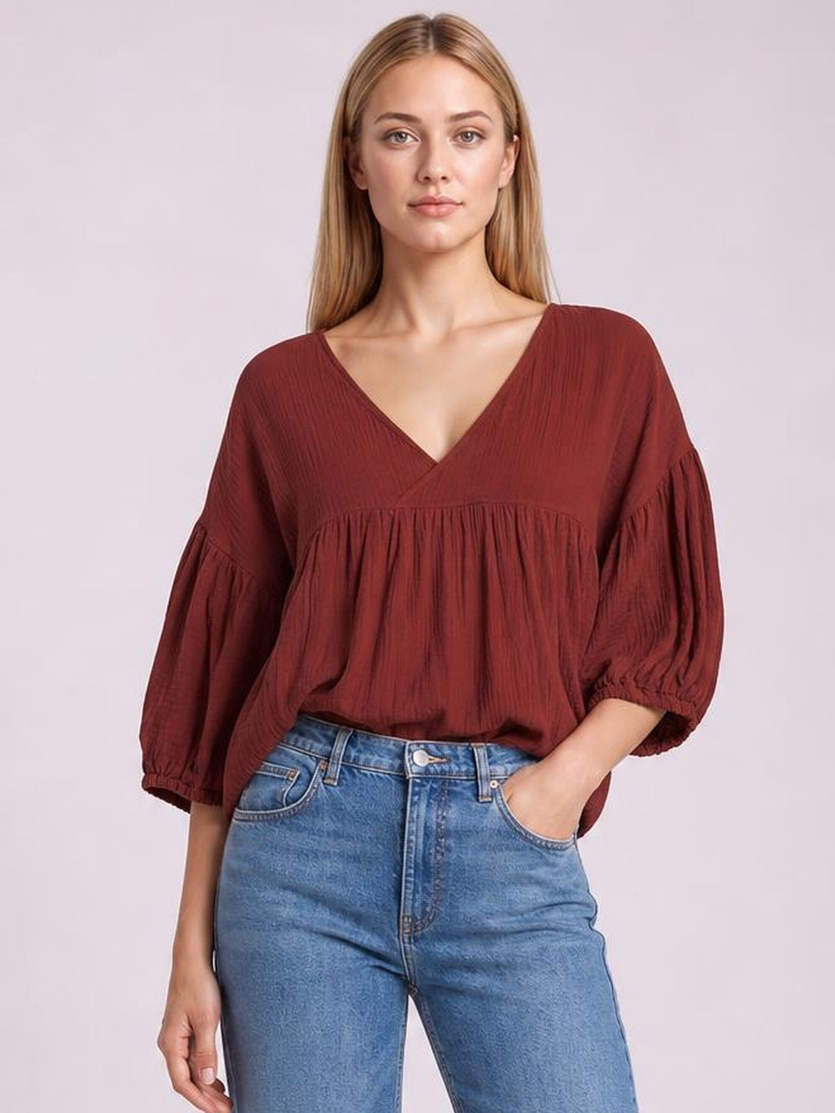 A model wears a solid rust-red dropped shoulder V-neck blouse with long gathered sleeves, elastic cuffs, and a relaxed silhouette, posing with one hand in her jeans pocket.