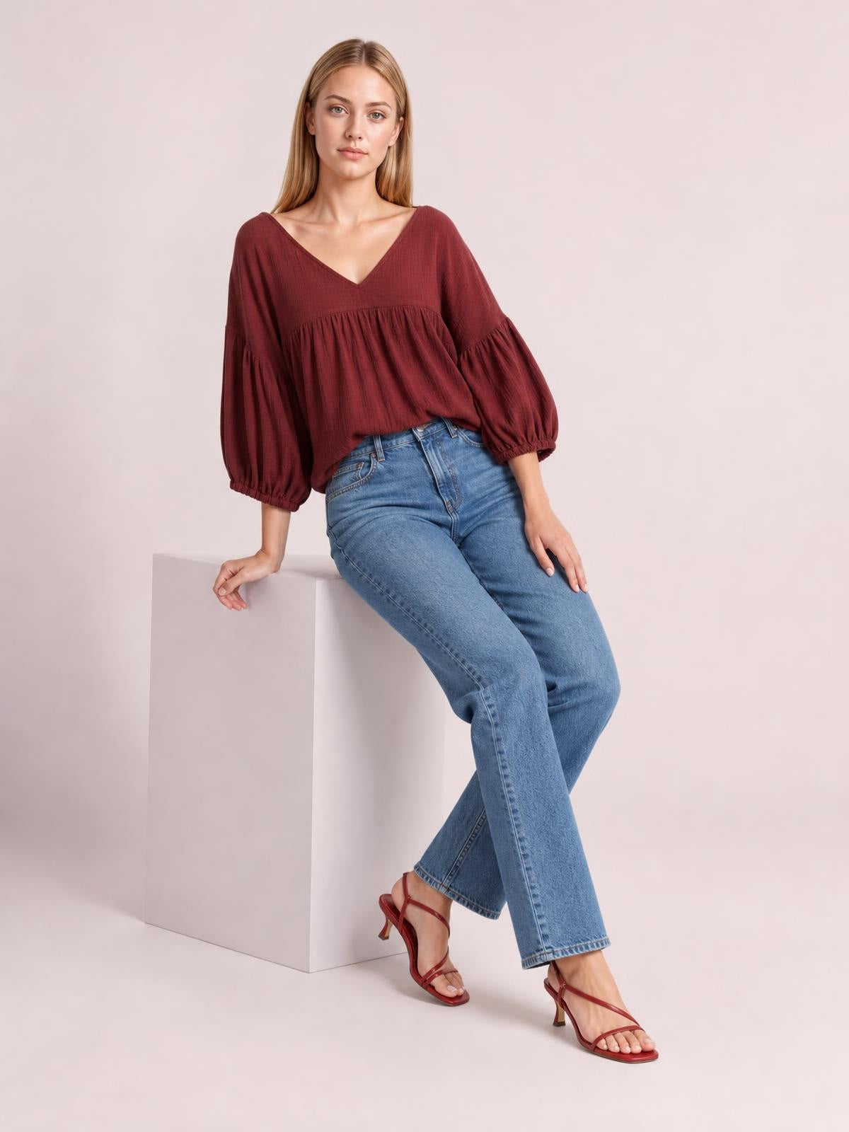 A model sits in a solid burgundy dropped-shoulder V-neck blouse with a relaxed long silhouette, gathered tiered body, and voluminous sleeves with elastic cuffs.