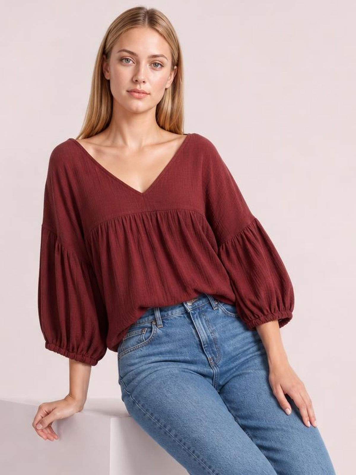A seated model wears a solid burgundy dropped shoulder V-neck blouse with long voluminous sleeves, gathered cuffs, and a relaxed silhouette.