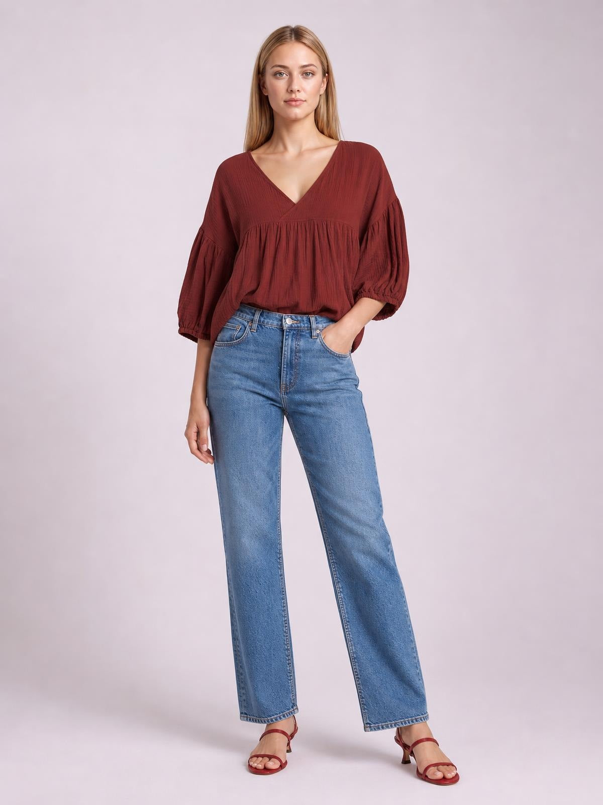 A model wears a solid burgundy V-neck blouse with dropped shoulders, long gathered sleeves, and a relaxed silhouette, posing with one hand in her jeans pocket.