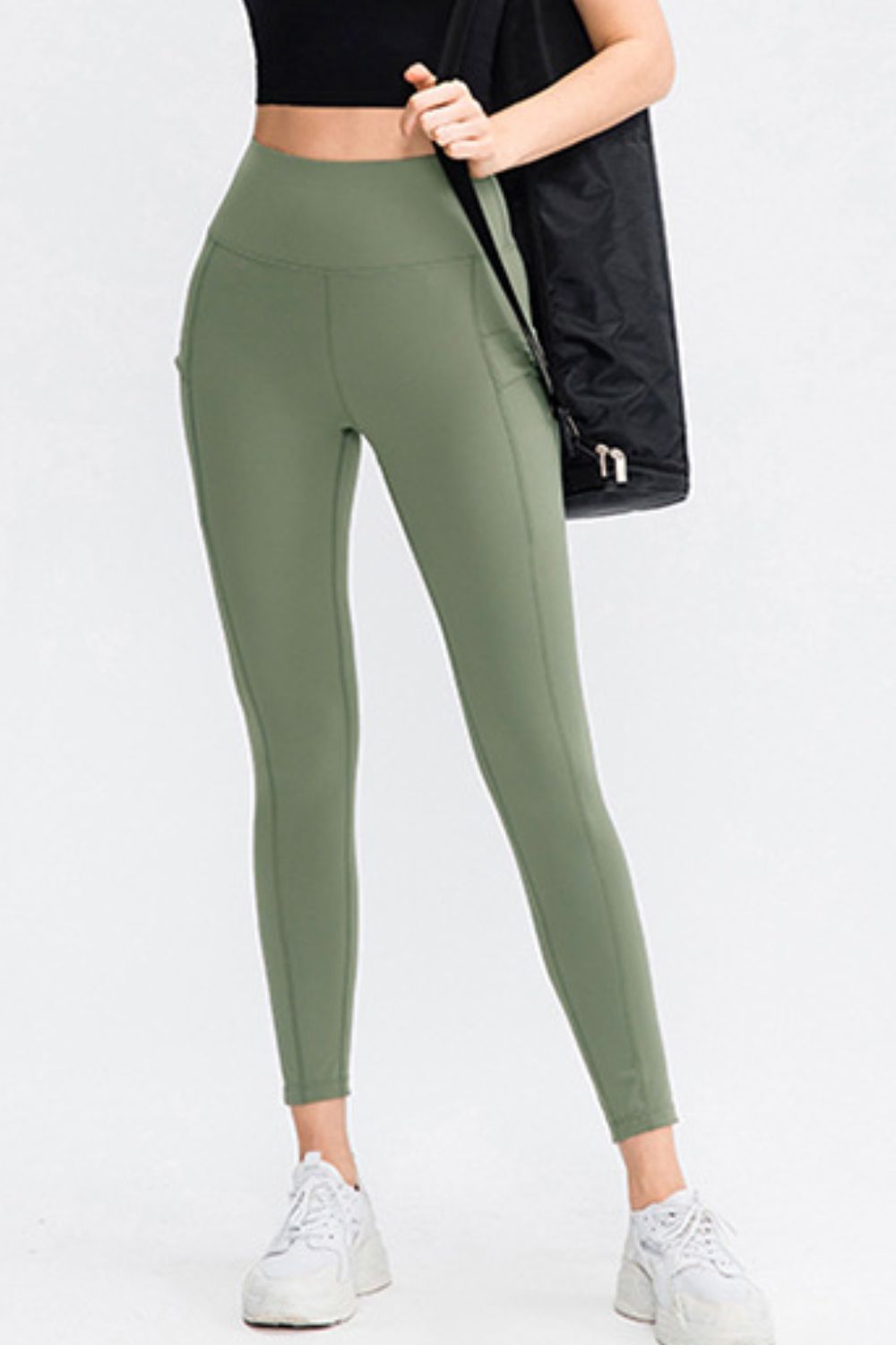 Model wearing solid olive-green slim-fit sports leggings with a wide waistband, long tapered silhouette, and side pocket, standing with one leg angled.