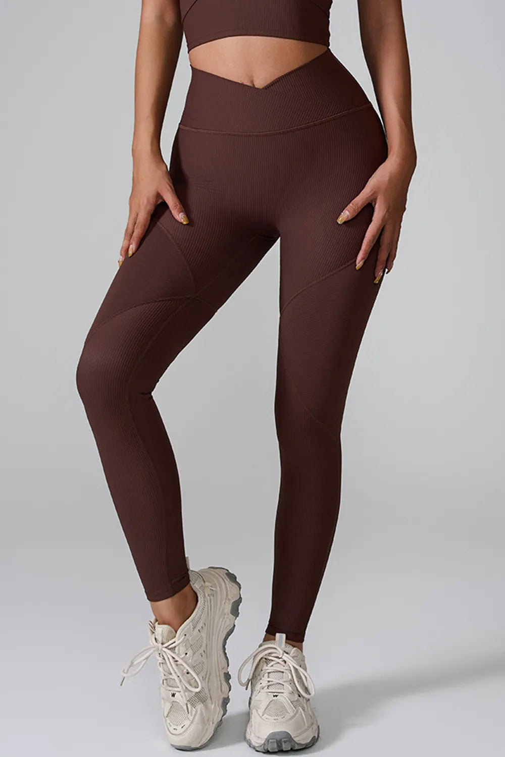Model wears high-waist active leggings in dark brown with a wide waistband, ribbed texture, curved seam panels, and a slim ankle-length silhouette.