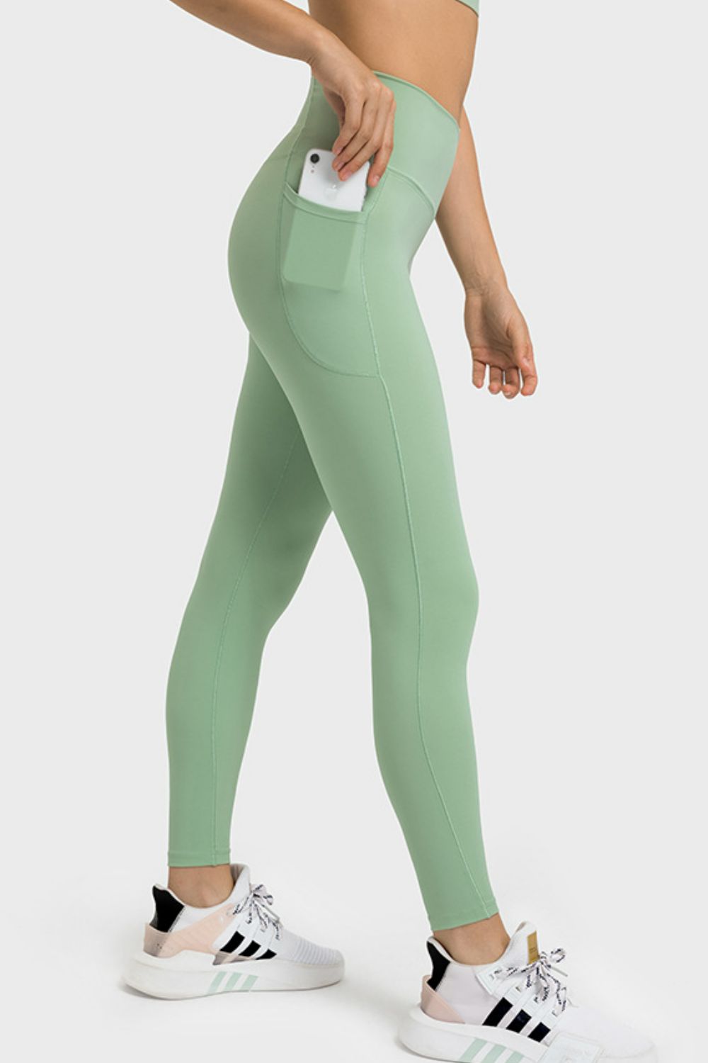 Model wears sage green V-waist yoga leggings with a fitted full-length silhouette, side pockets, and visible seam construction while standing in a walking pose.