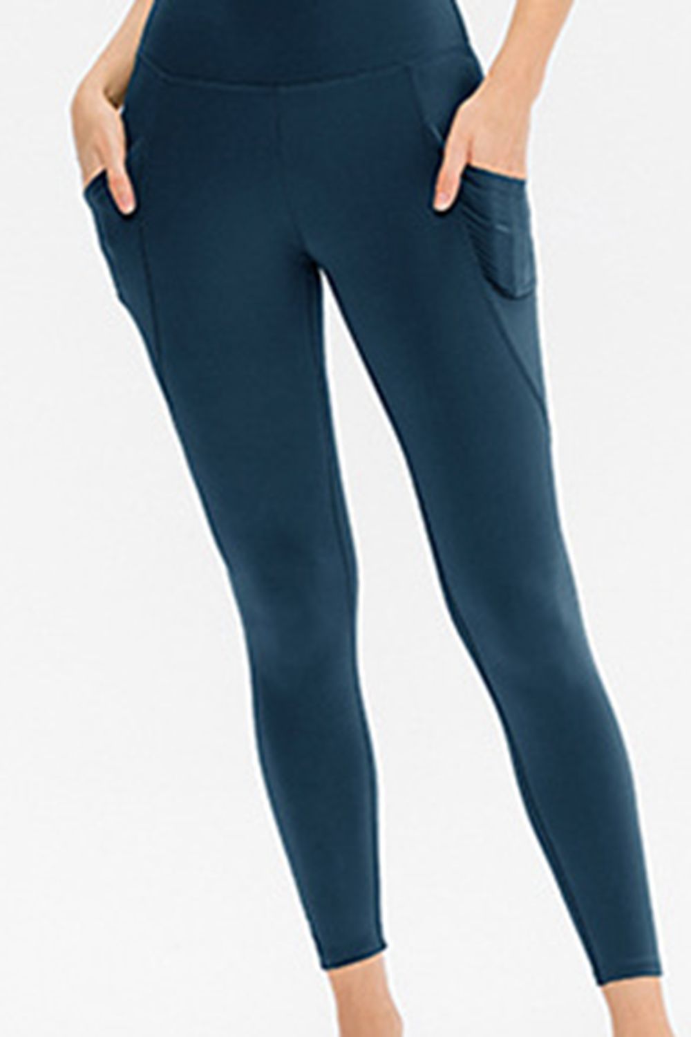 A model wears solid navy slim-fit long active leggings with a smooth waistband, ankle-length silhouette, and side pockets, standing with hands tucked inside.