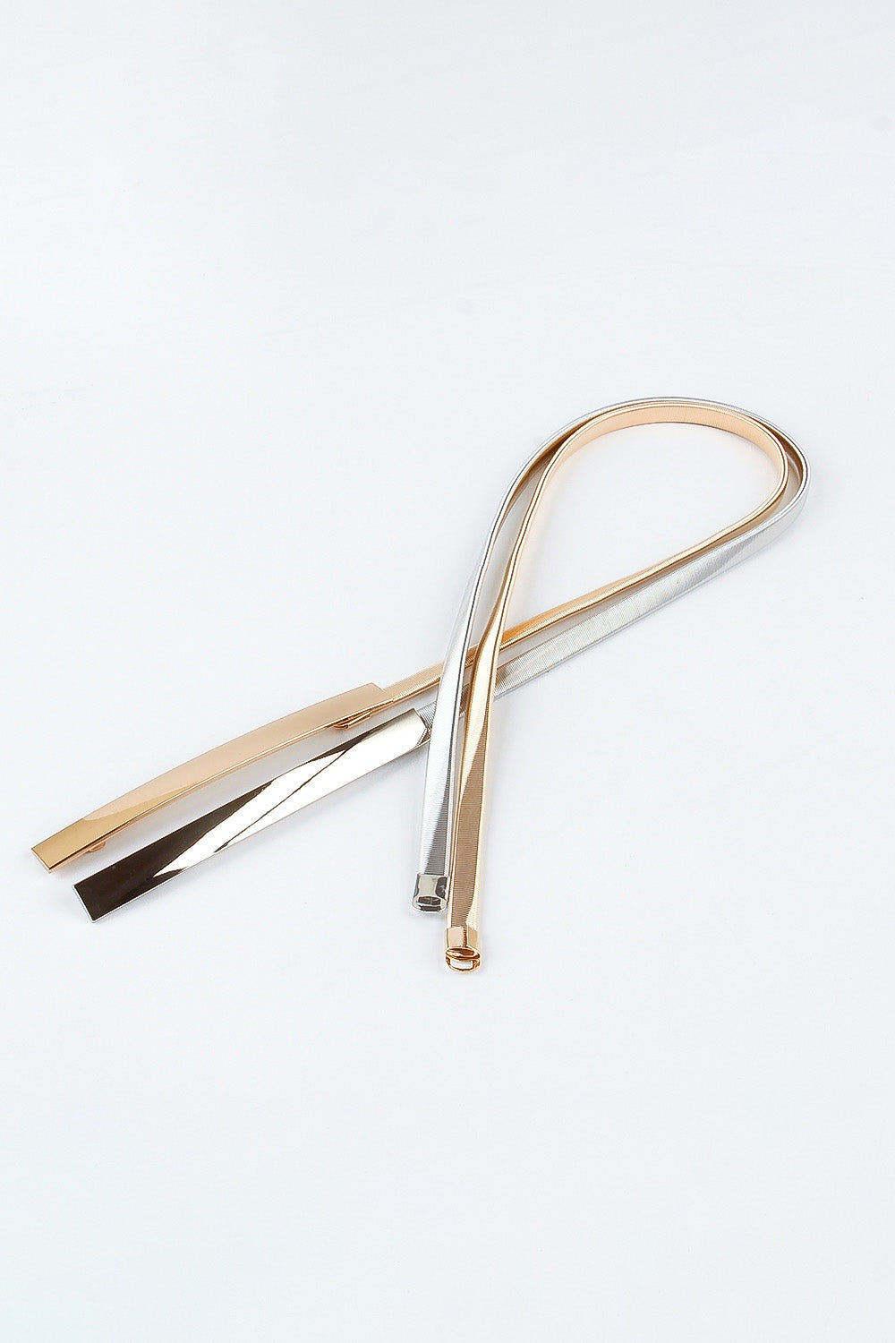 Several sleek iron skinny belts in polished gold, silver, and black finishes are arranged in overlapping loops, showing their slender construction and fastening ends.