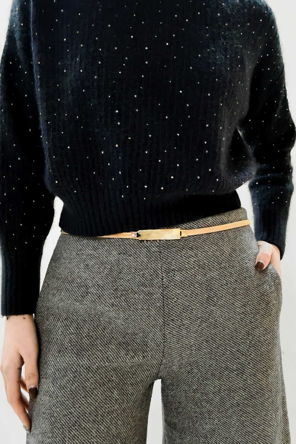 A cropped model wears a slender gold-tone elastic iron skinny belt with a structured rectangular buckle over gray tailored trousers and a black embellished sweater.
