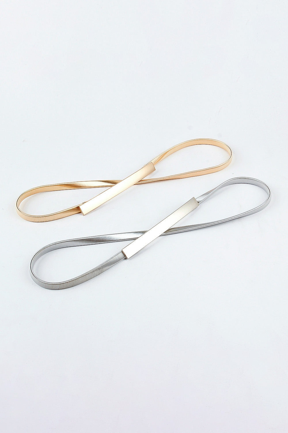 Two slim elastic iron belts in gold and silver tones are displayed in loops, each finished with a clean rectangular buckle and fine ribbed construction.