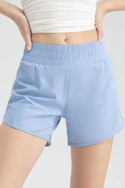 Model wearing light blue elastic waist active shorts with a relaxed silhouette, wide gathered waistband, curved hems, and hands resting at the hips.