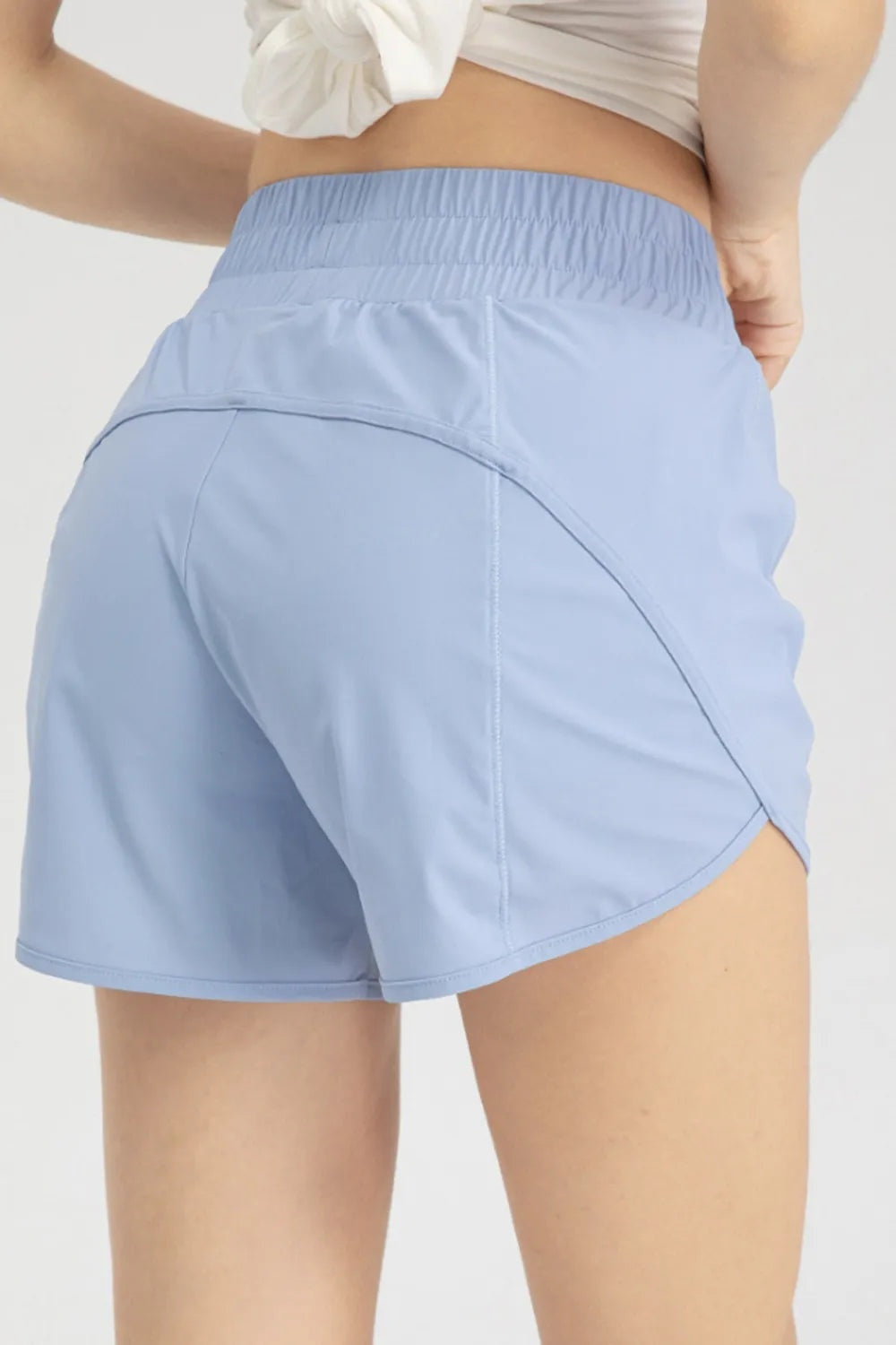 Model shown from behind wears light blue elastic waist active shorts with a relaxed short silhouette, curved panel seams, and neatly finished hems.
