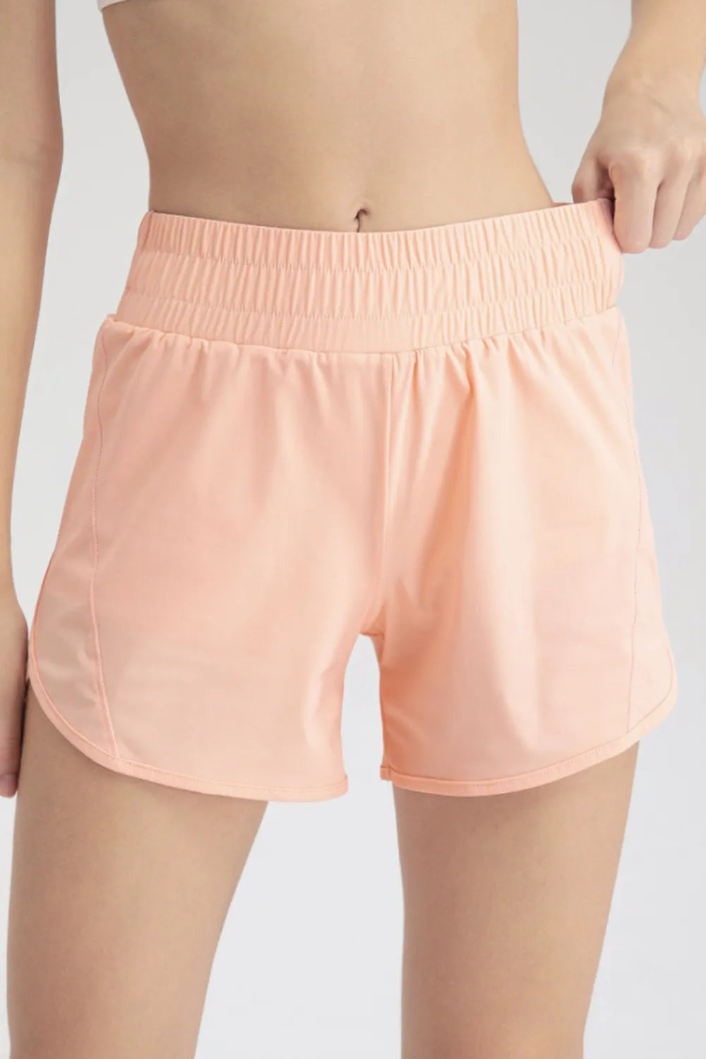 Model wears peach elastic waist active shorts with a relaxed, curved-leg silhouette, gathered waistband, and neatly stitched hems.