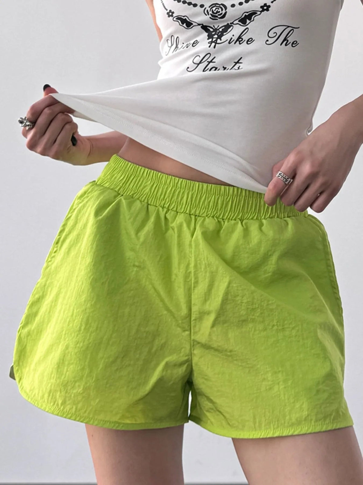 Model lifts a white top to show lime green active shorts with an elastic waist, relaxed loose silhouette, lightweight crinkled construction, and curved hems.