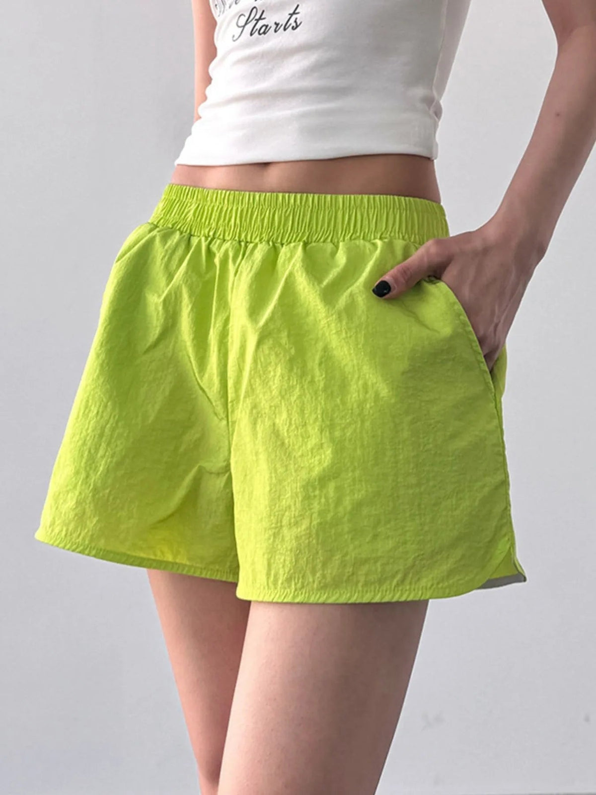 Model wearing lime green elastic waist active shorts with a lightweight relaxed silhouette, gathered waistband, and hand resting in the side pocket.