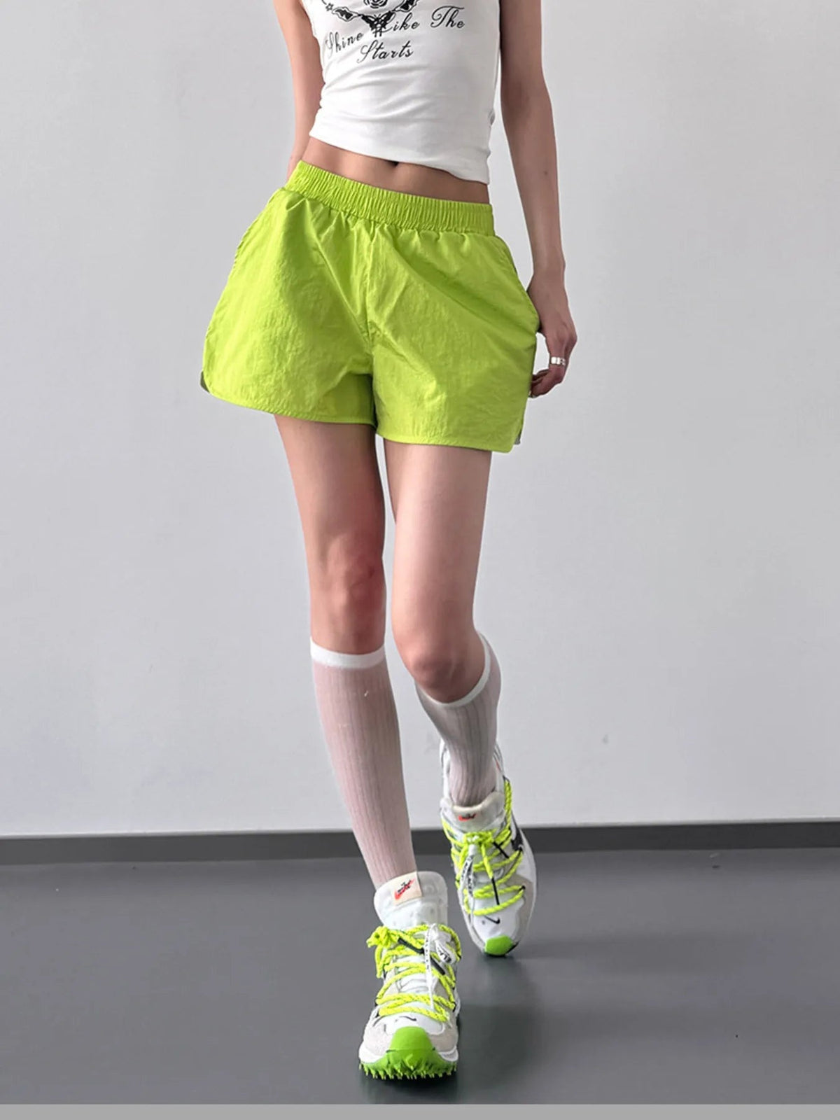 Model wearing bright lime green elastic waist active shorts with a lightweight, relaxed fit and straight hems, standing with one leg crossed forward.