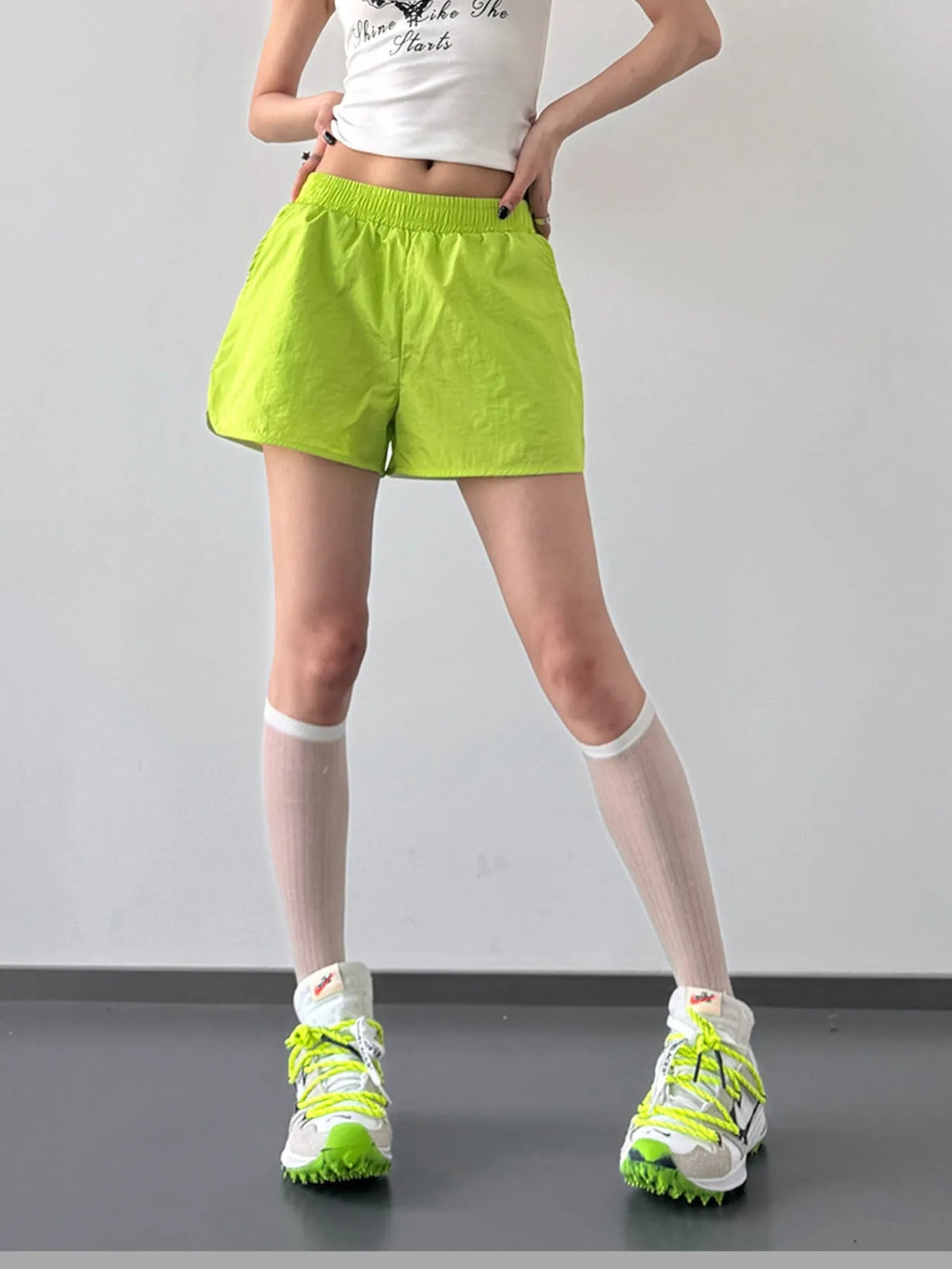 Model poses with hands at the waistband of bright lime green elastic waist active shorts, featuring a lightweight loose silhouette and clean hem.