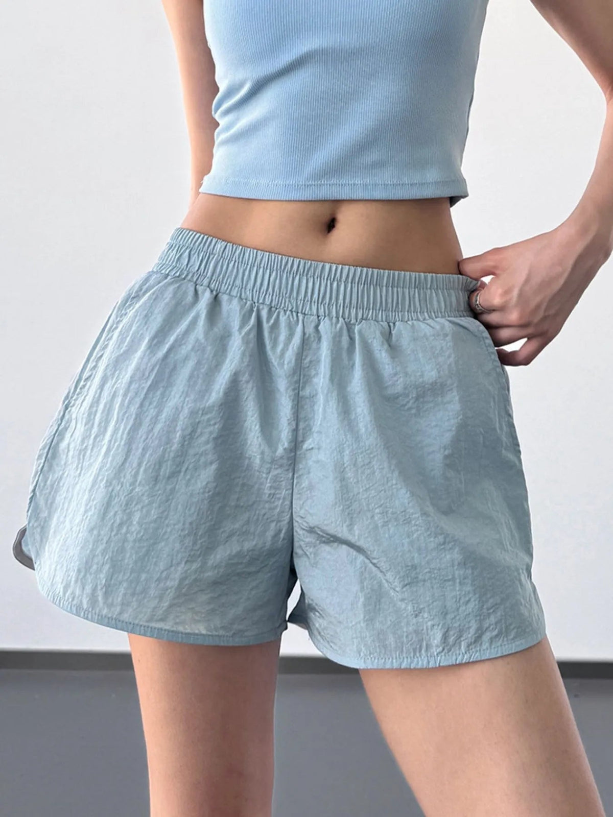 Model wears light blue elastic waist active shorts with a relaxed, lightweight silhouette, wide waistband, and straight hems while adjusting the waistband.