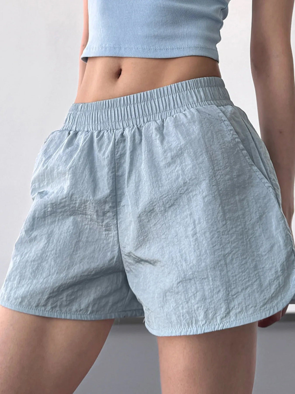 A model stands wearing light blue Elastic Waist Active Shorts with a gathered elastic waistband, relaxed loose silhouette, and crinkled lightweight fabric.