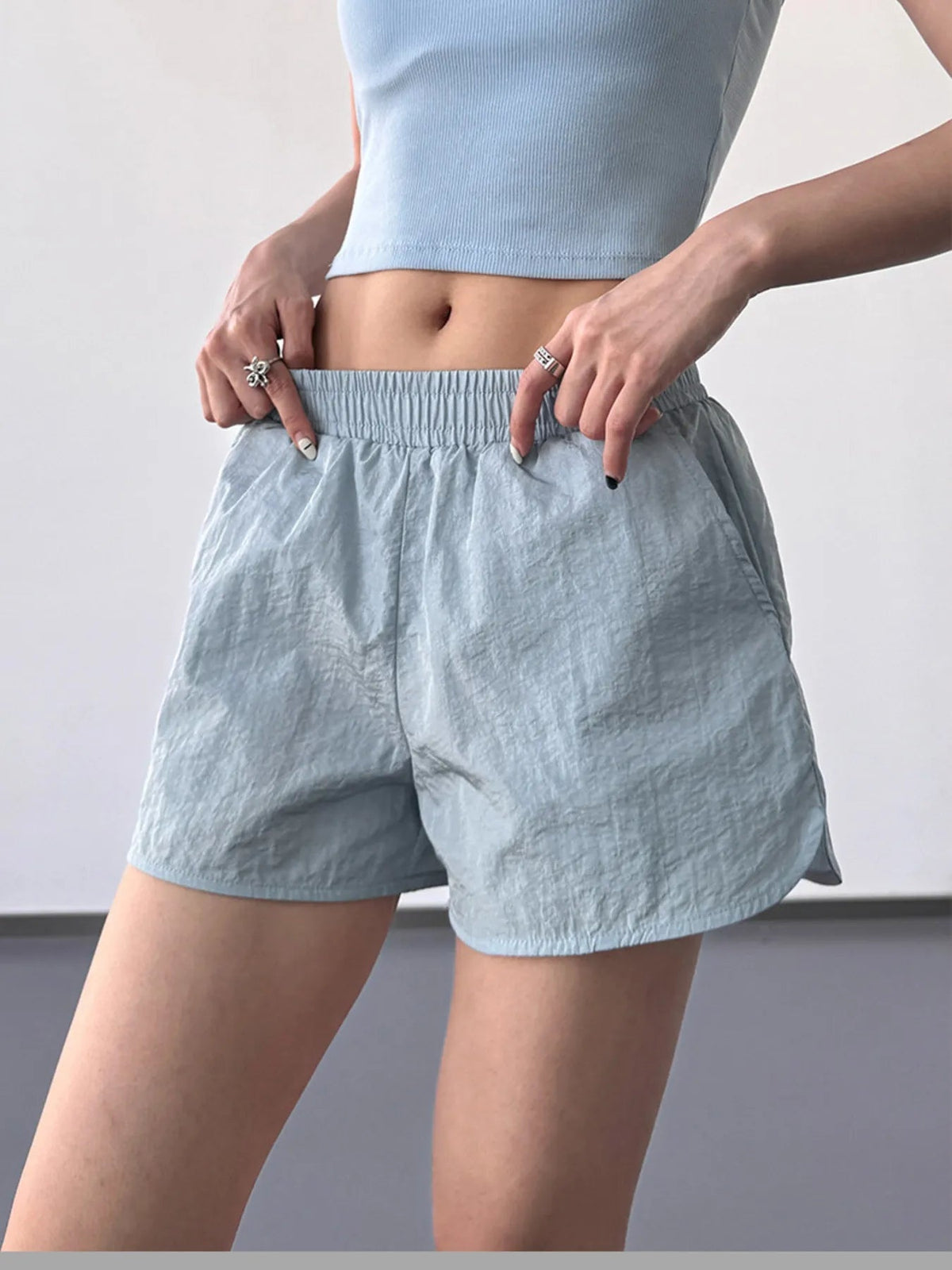 Model points to light blue elastic waist active shorts with a relaxed, lightweight silhouette, wide waistband, and softly crinkled fabric.