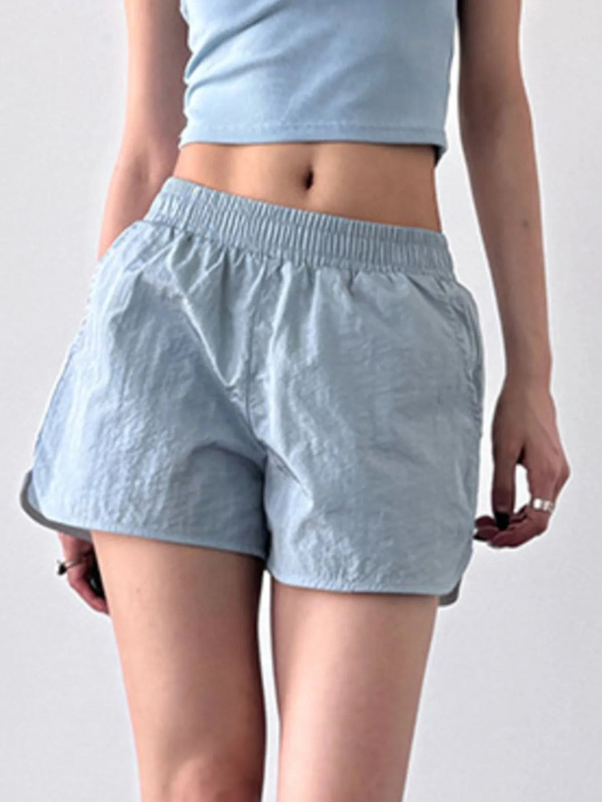 A model stands in light blue elastic-waist active shorts with a relaxed, loose silhouette, curved hems, and lightweight crinkled fabric.