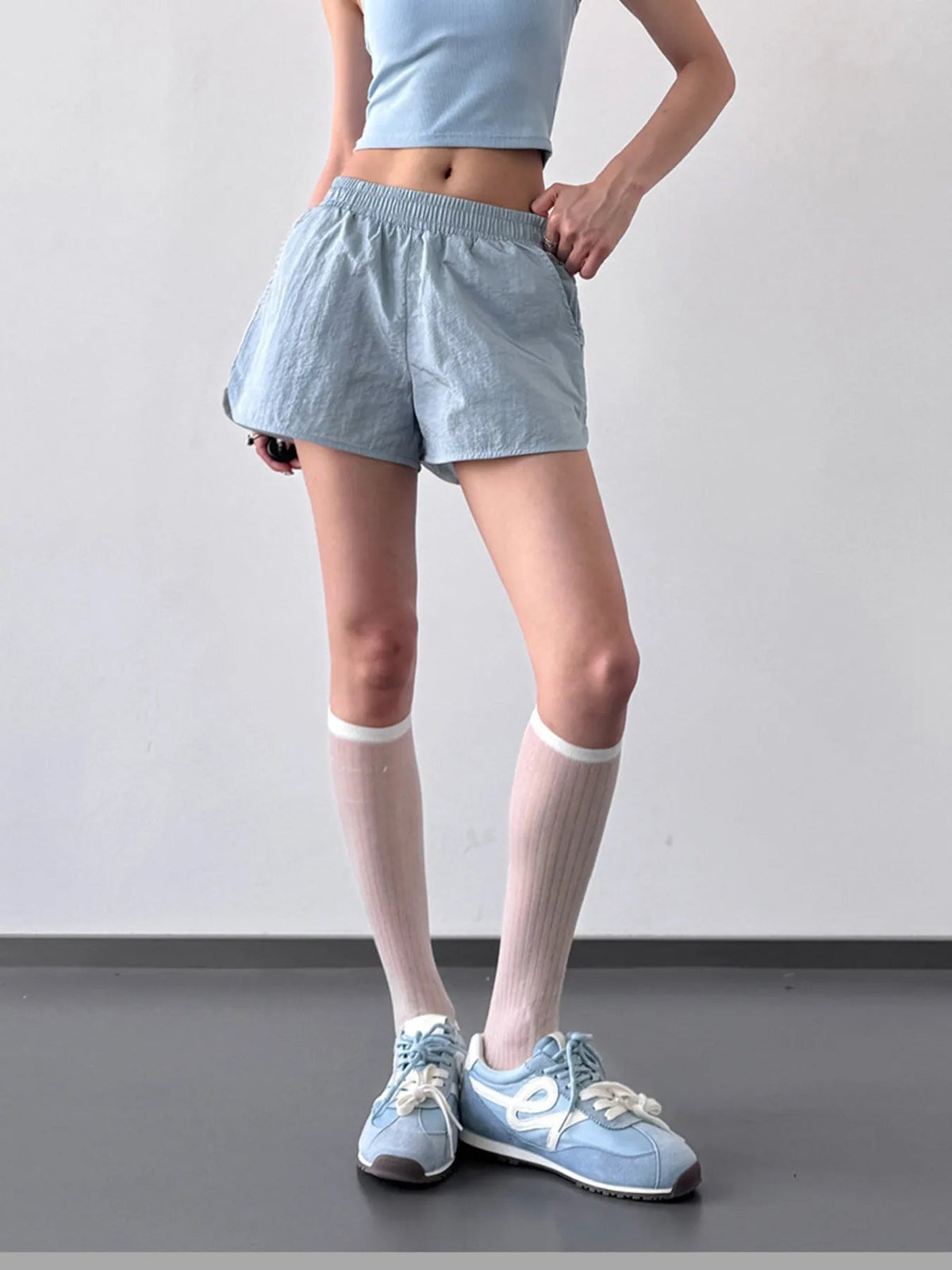 Model wearing light blue Elastic Waist Active Shorts with a gathered elastic waistband, lightweight crinkled fabric, and a relaxed short silhouette while standing with one hand at the waist.