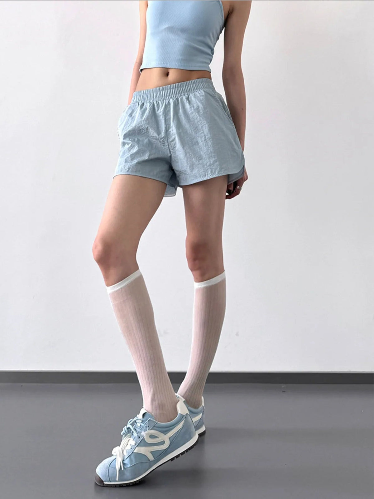 Model wears light blue elastic waist active shorts with a relaxed, lightweight silhouette and curved hems, posing with one leg crossed forward.