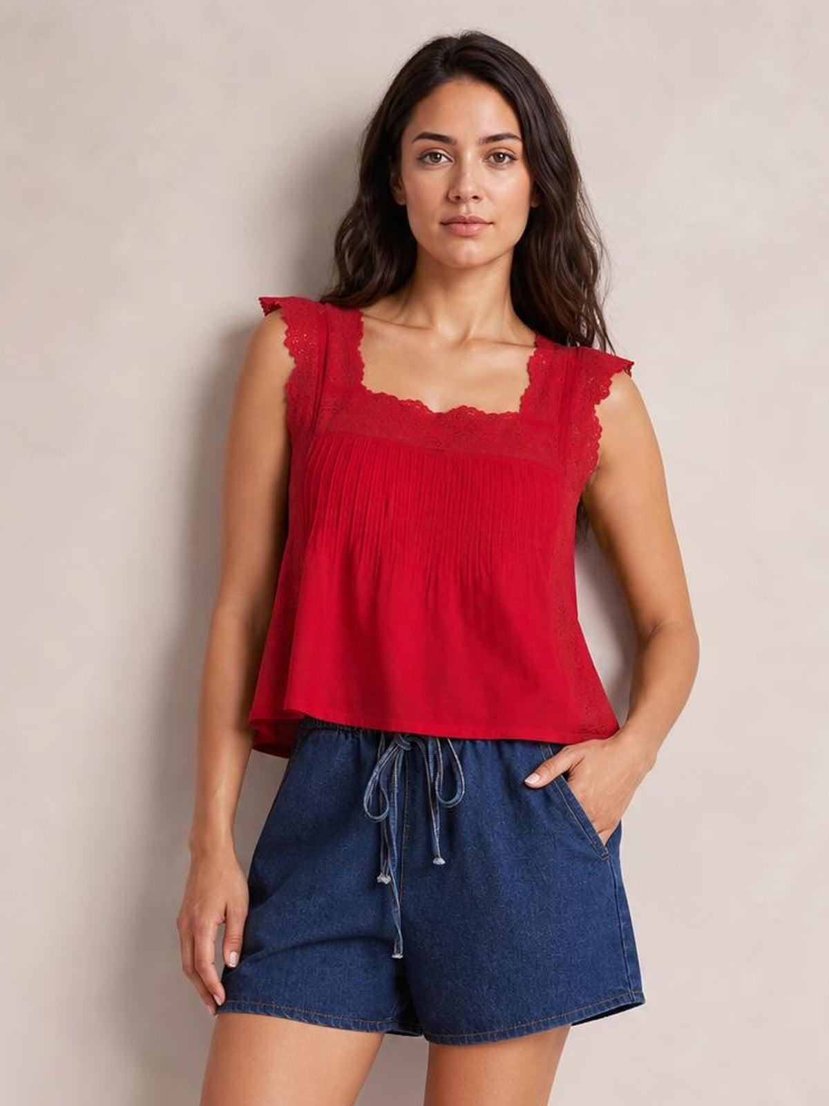 A woman modeling a sleeveless red top with a square neckline, delicate lace trim, and a smocked bodice, paired with blue denim shorts against a neutral beige background.