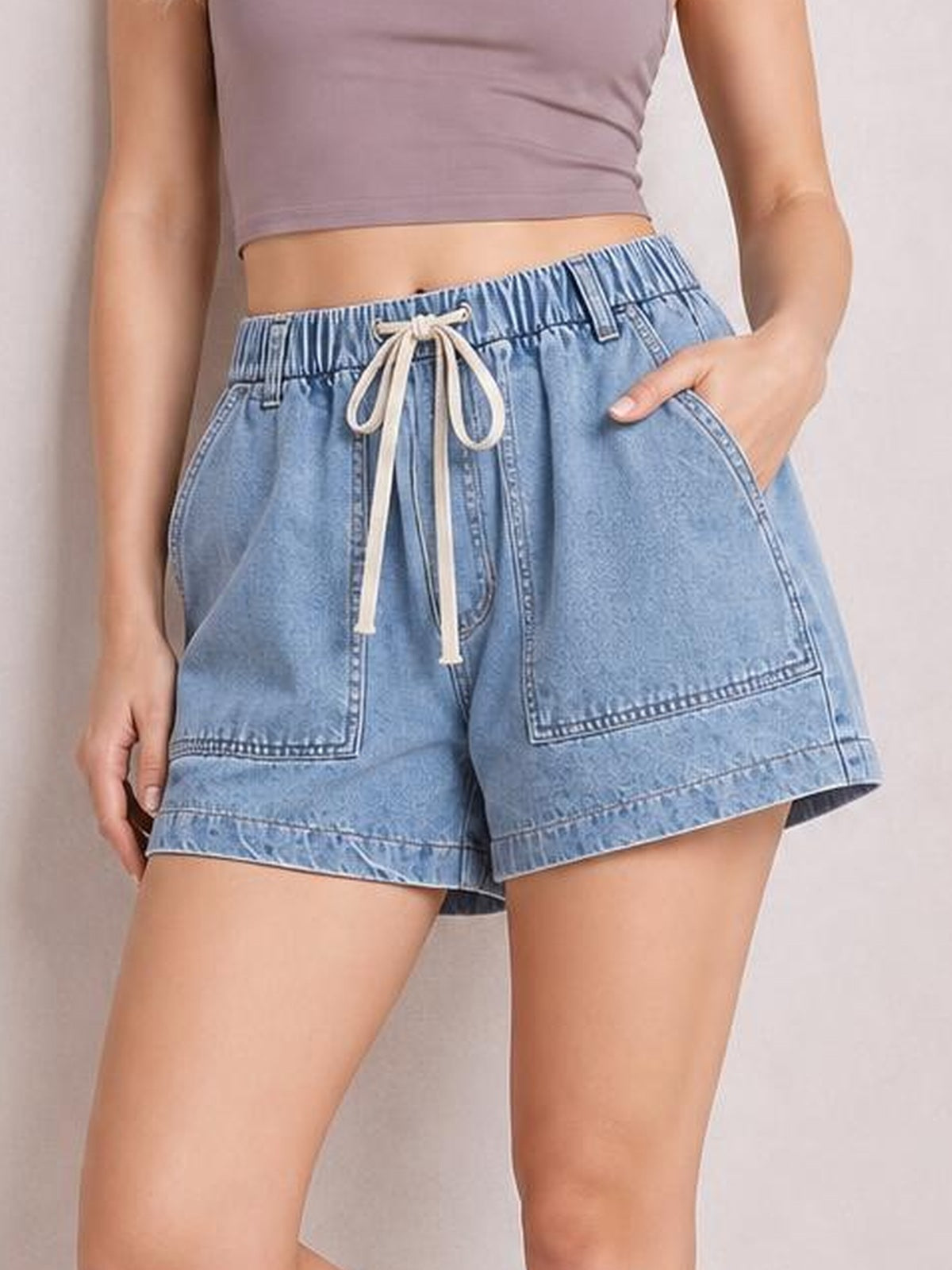 A model wears light blue denim shorts with an elastic drawstring waist, roomy pocketed construction, stitched panel details, and a relaxed straight-leg silhouette.