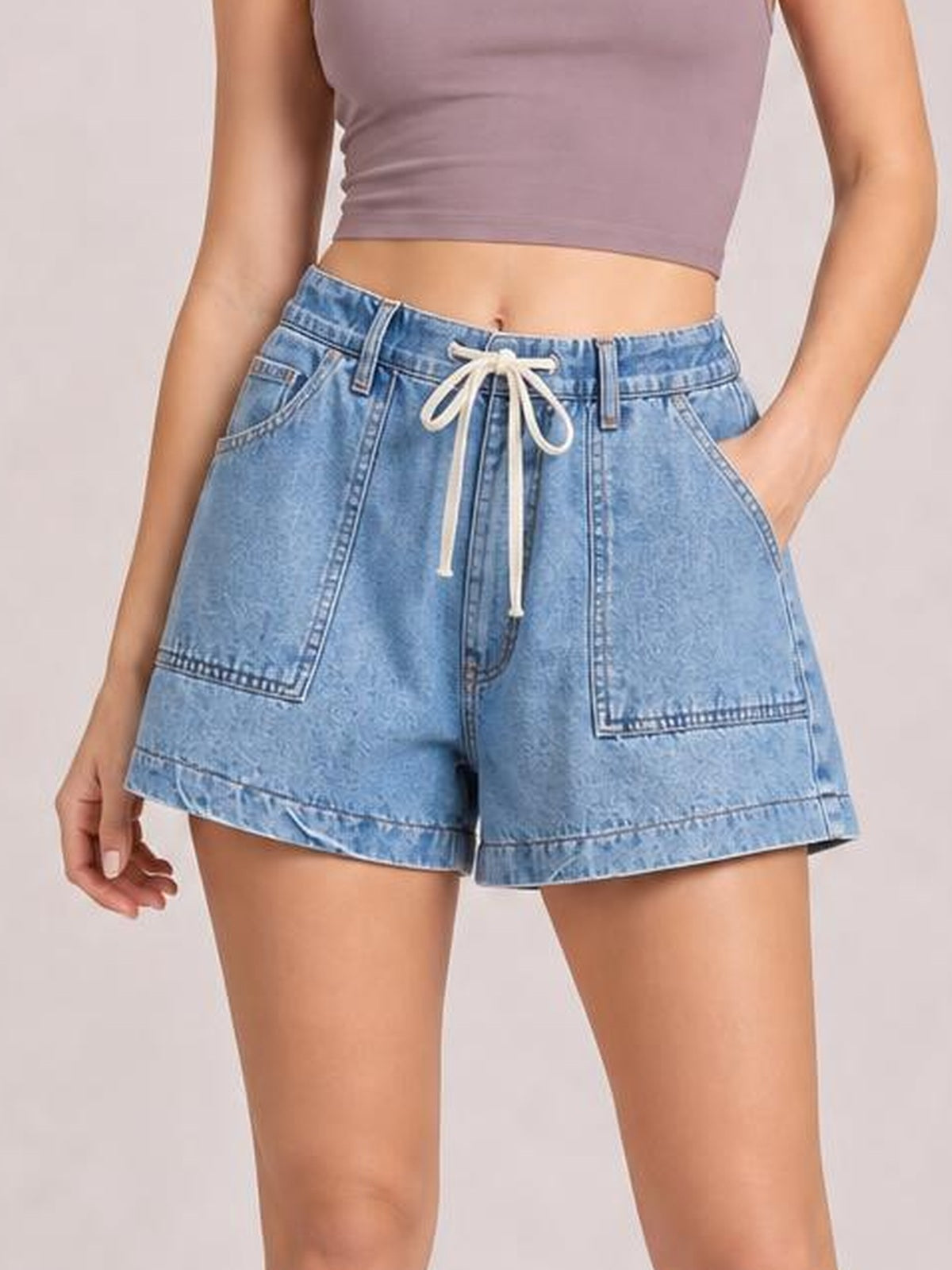 A standing model wears light blue elastic waist denim shorts with a drawstring, relaxed silhouette, large front pockets, and stitched straight hems.