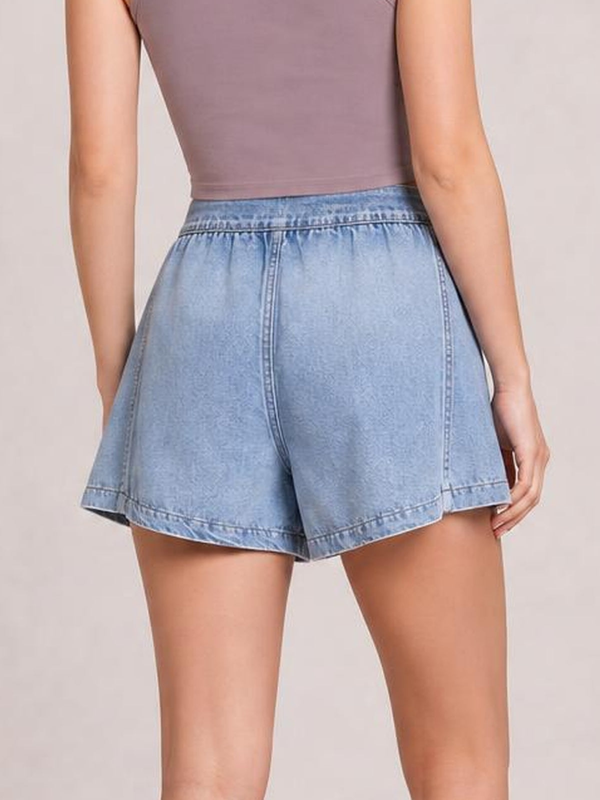 A back view shows a model wearing light blue elastic waist denim shorts with a relaxed, flared silhouette, curved hem, and visible seam detailing.
