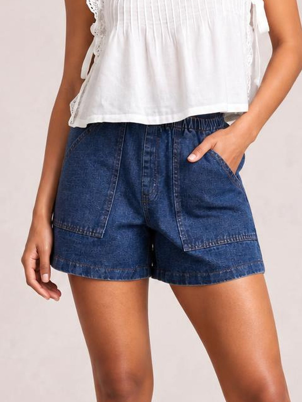 Model wearing dark blue elastic waist denim shorts with a drawstring, relaxed straight-leg silhouette, front patch pockets, and stitched hems.