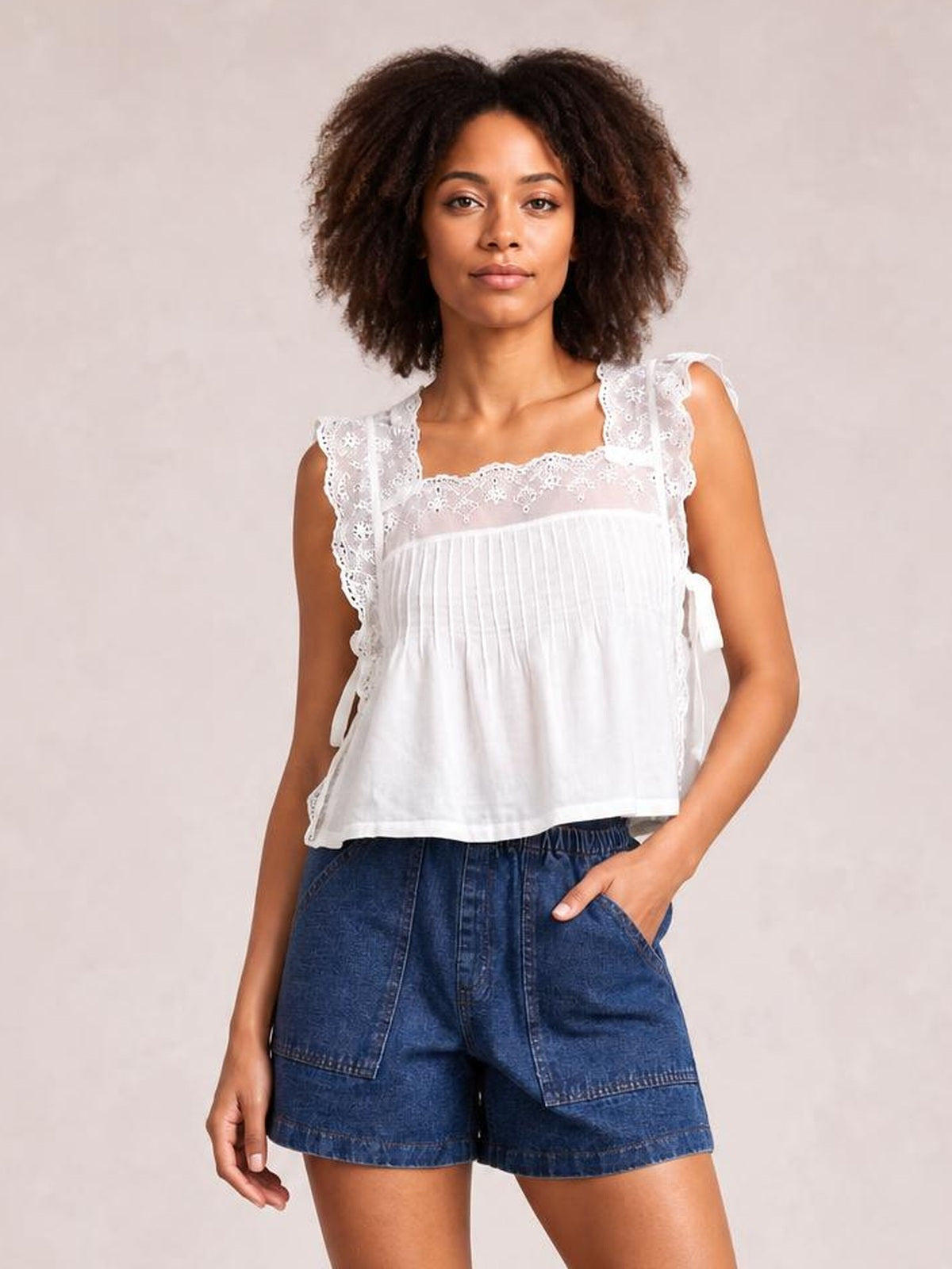 A woman modeling a white sleeveless top with delicate lace trim and ruffle details, paired with blue denim shorts, against a neutral beige background.