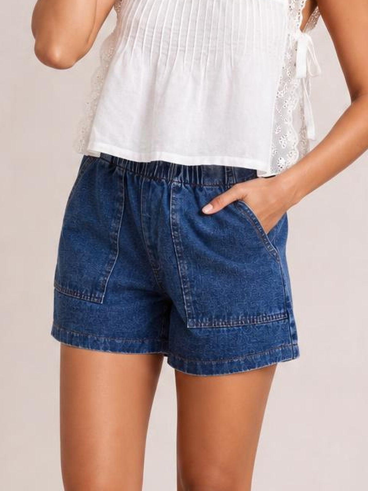 Model wearing dark blue elastic waist denim shorts with a drawstring, roomy pocketed design, relaxed silhouette, and straight hems, with one hand in a pocket.