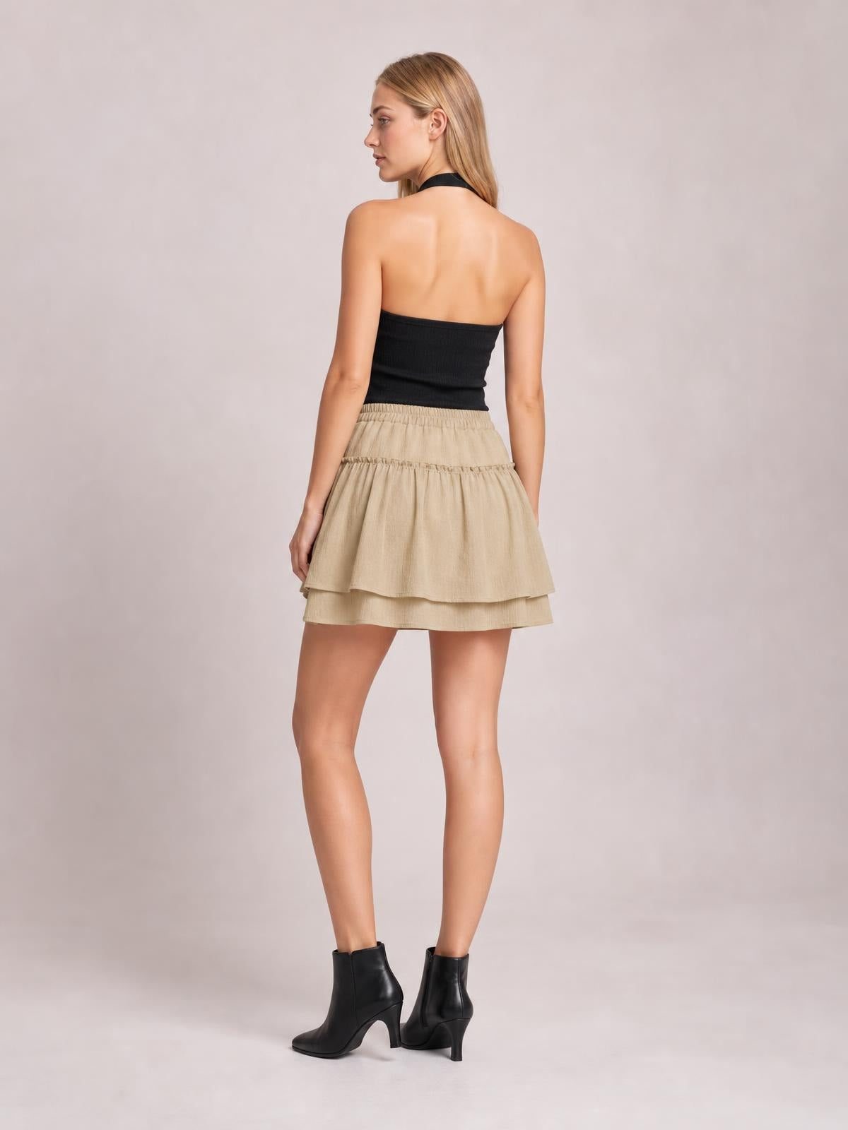 A model poses from the back in a beige elastic waist layered skirt with a flowy short silhouette, gathered tiers, and frill detailing.