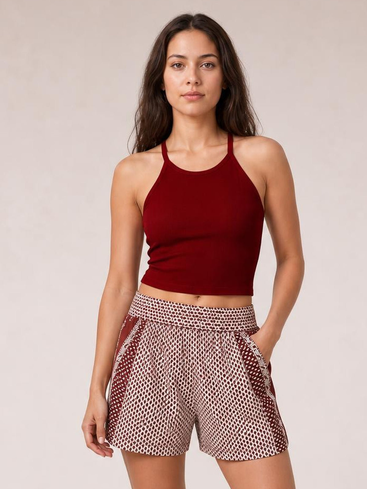 A woman modeling a dark red ribbed halter crop top and patterned shorts with a red and white geometric print, set against a plain beige studio background.