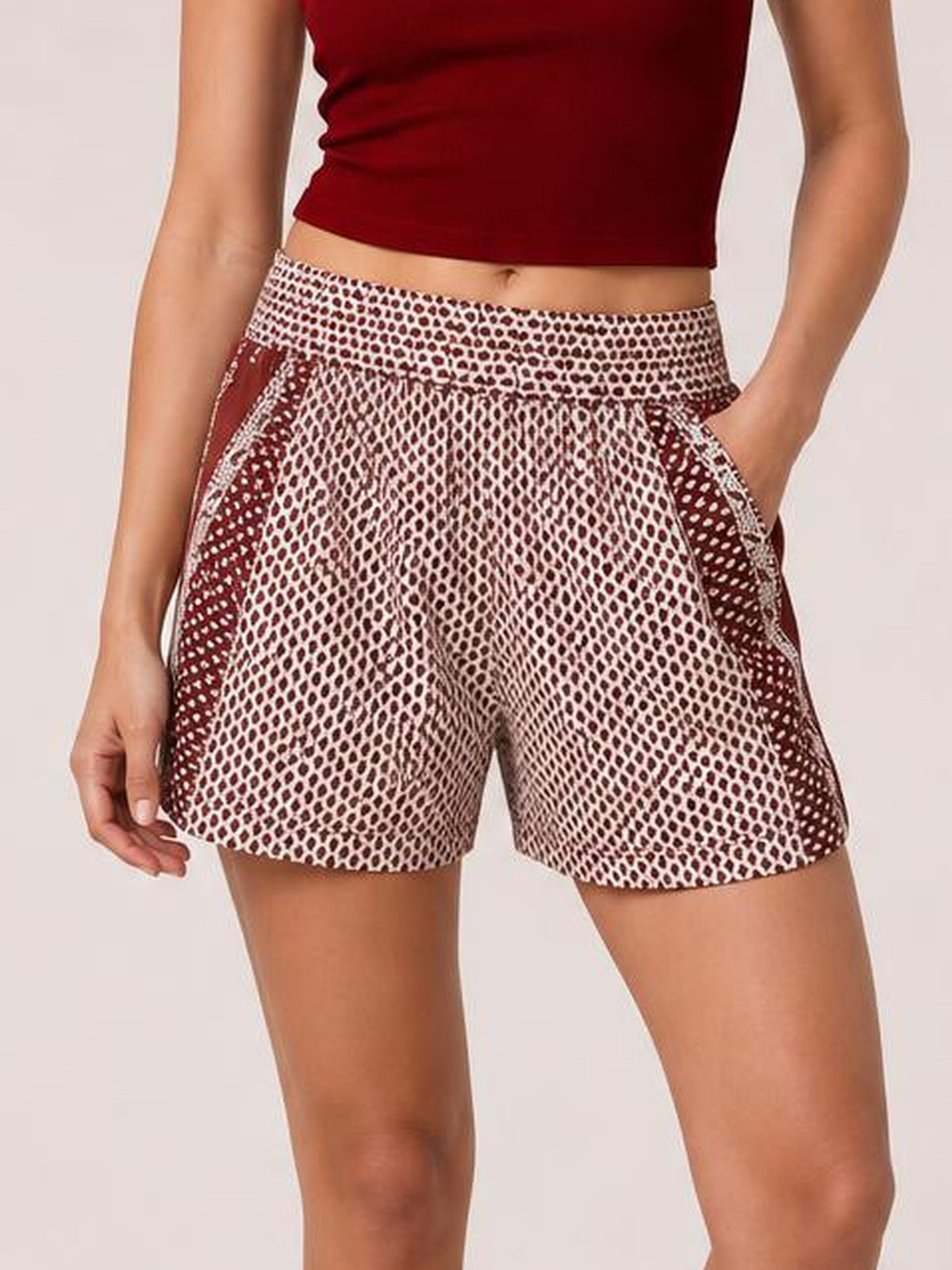 A model stands wearing burgundy and white patterned elastic waist shorts with a relaxed silhouette, side pockets, and one hand tucked into a pocket.