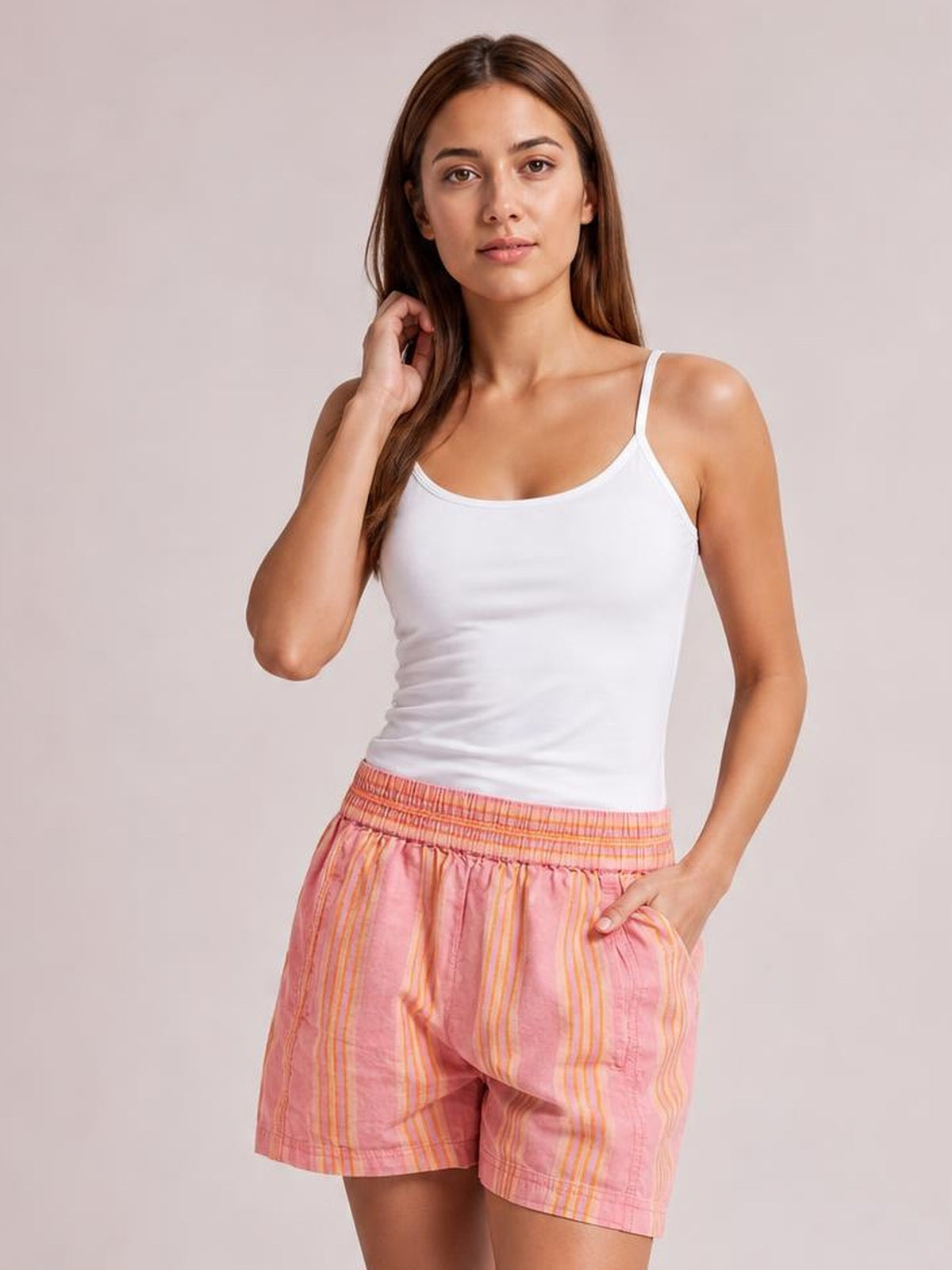 A woman modeling a white ribbed tank top and pink striped shorts with orange and yellow vertical lines, set against a solid light beige studio background.