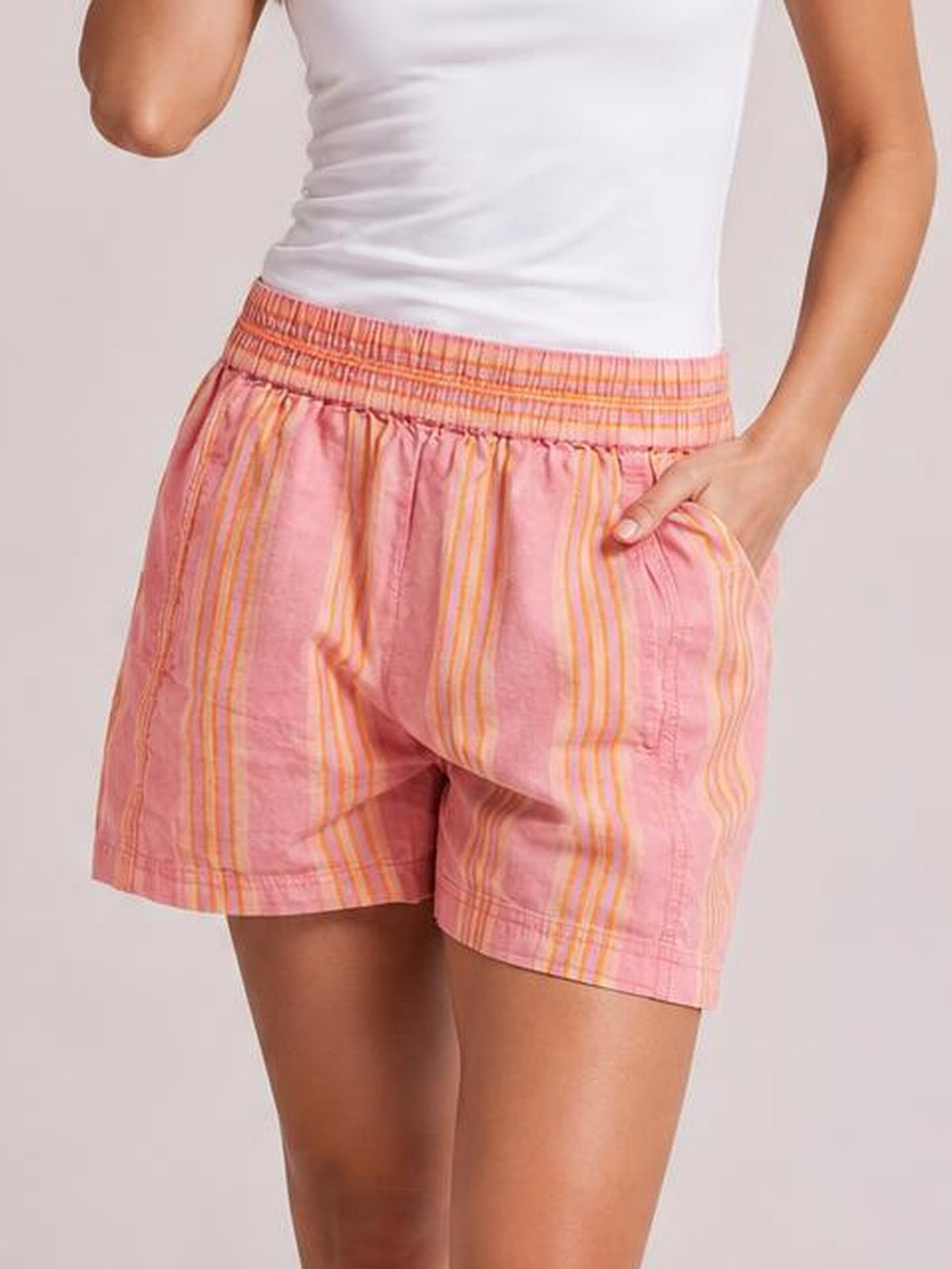 Model wears pink striped elastic waist shorts with a relaxed silhouette, side pockets, and neatly hemmed legs, shown with one hand in a pocket.