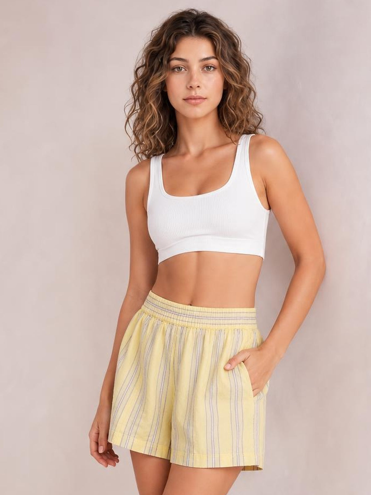 A woman modeling a white scoop-neck crop top and yellow-and-white vertically striped shorts against a soft beige studio background.