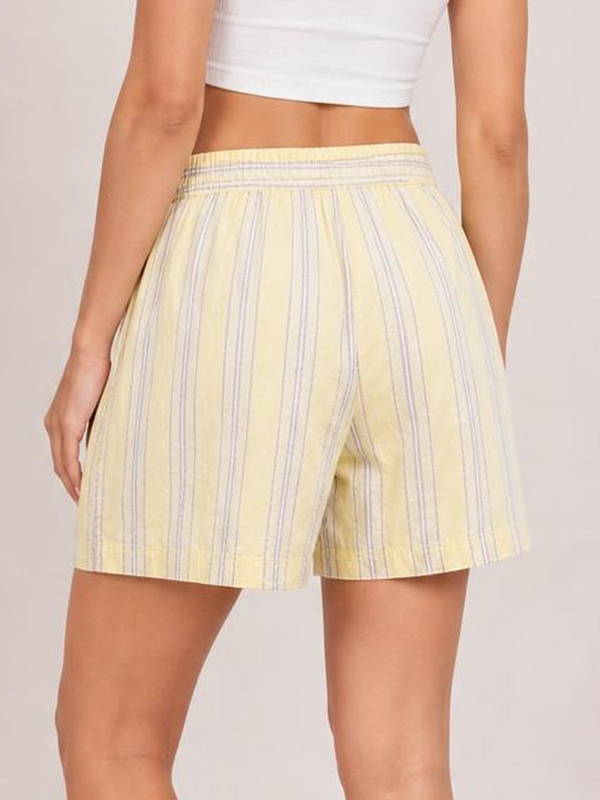 Model shown from behind wearing pale yellow elastic waist shorts with vertical white and lavender stripes, a relaxed fit, and side pockets.