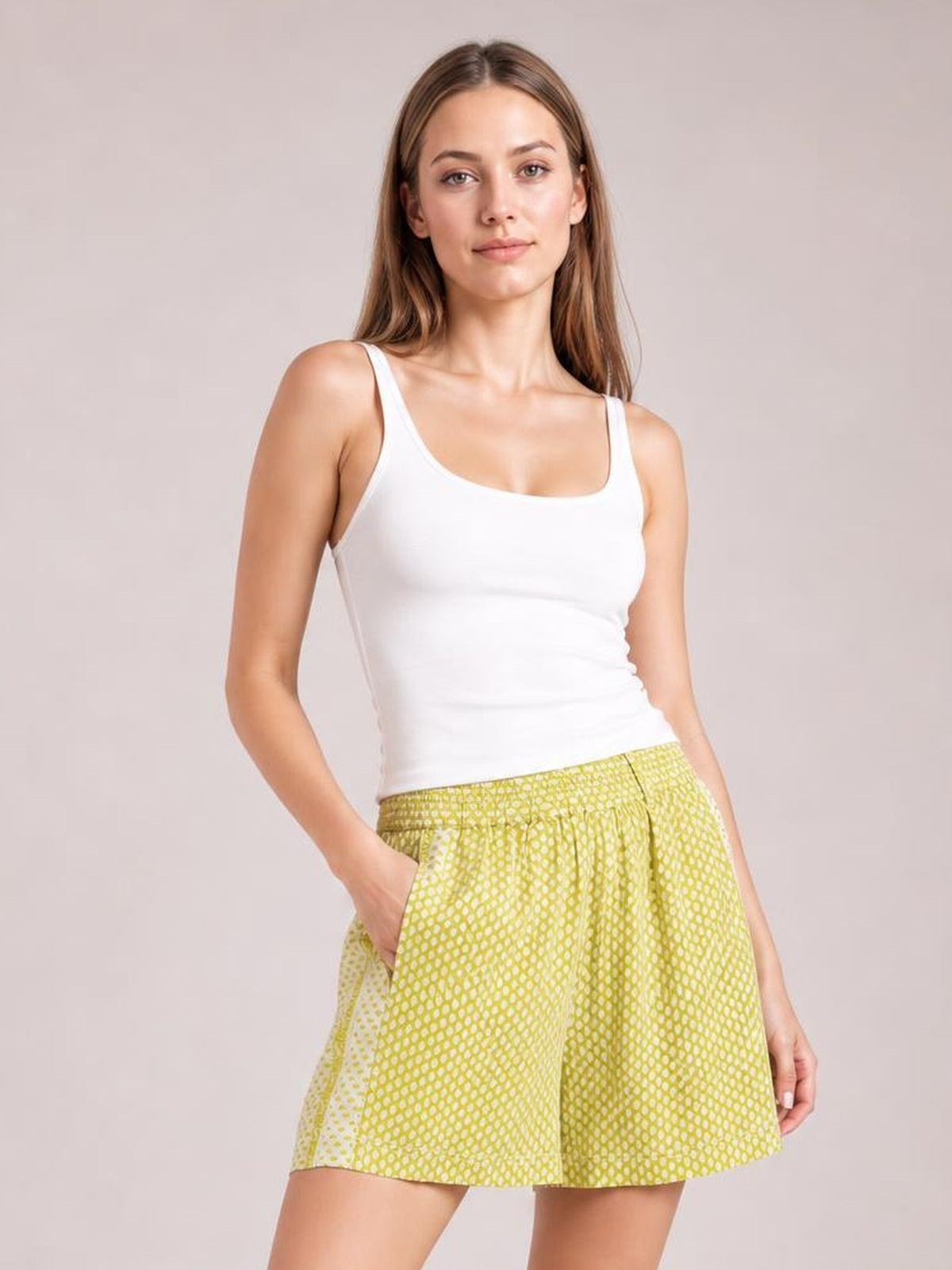 A woman modeling a white ribbed tank top and yellow-and-white patterned shorts against a solid light beige studio background.