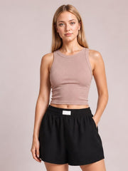 A woman modeling a sleeveless ribbed crop top in dusty rose and black athletic shorts, standing against a solid light beige studio background.