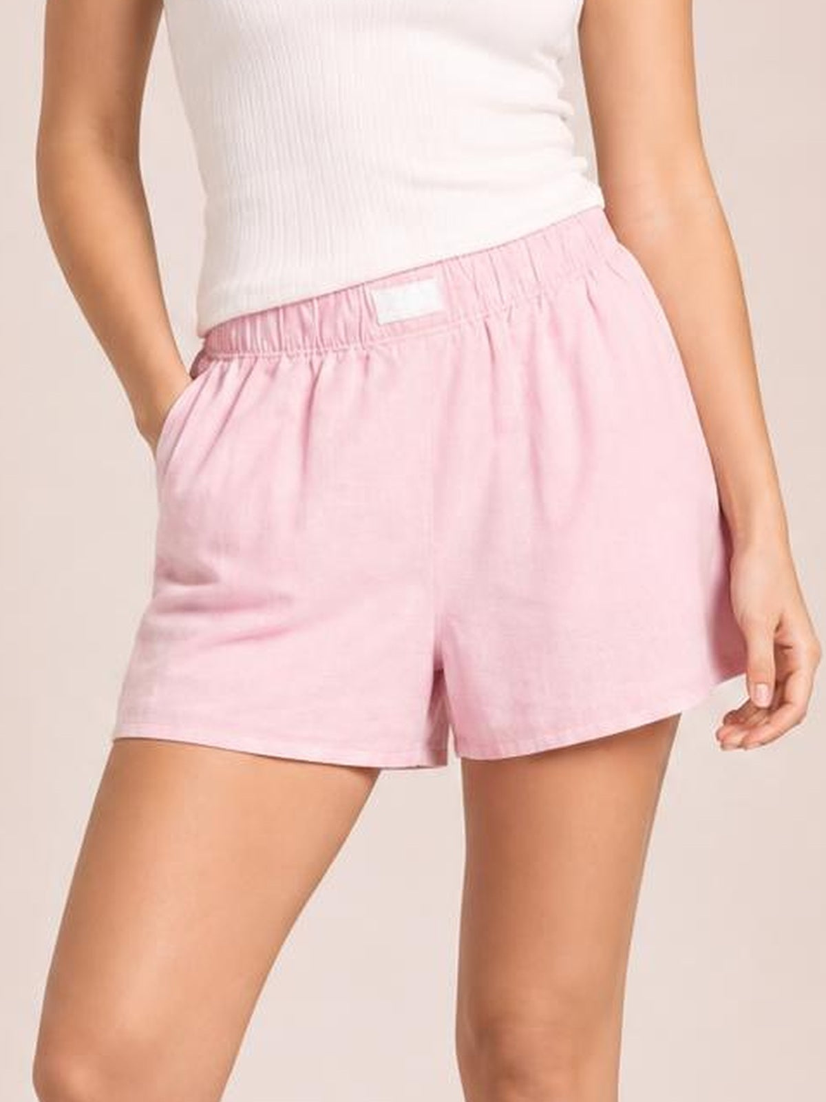 Model wears pink elastic waist shorts with a relaxed straight-leg silhouette, side pockets, and clean construction, posing with one hand tucked into a pocket.