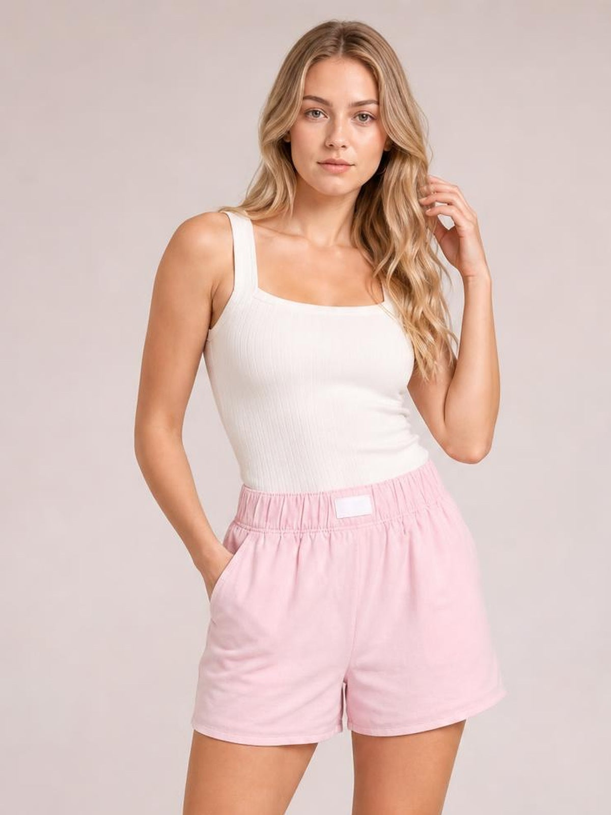A woman modeling a white ribbed tank top and light pink shorts against a solid light beige studio background.