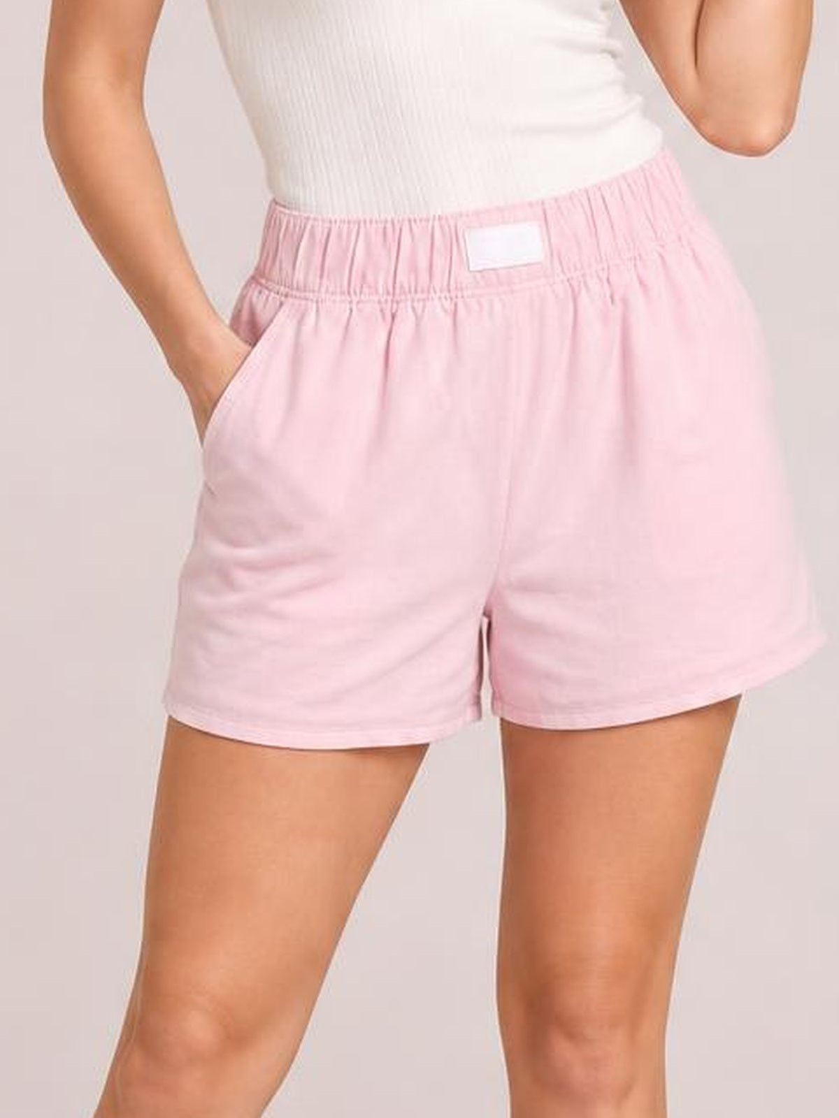 A model wears light pink elastic waist shorts with a relaxed silhouette, straight hems, and side pockets, posing with one hand tucked into a pocket.