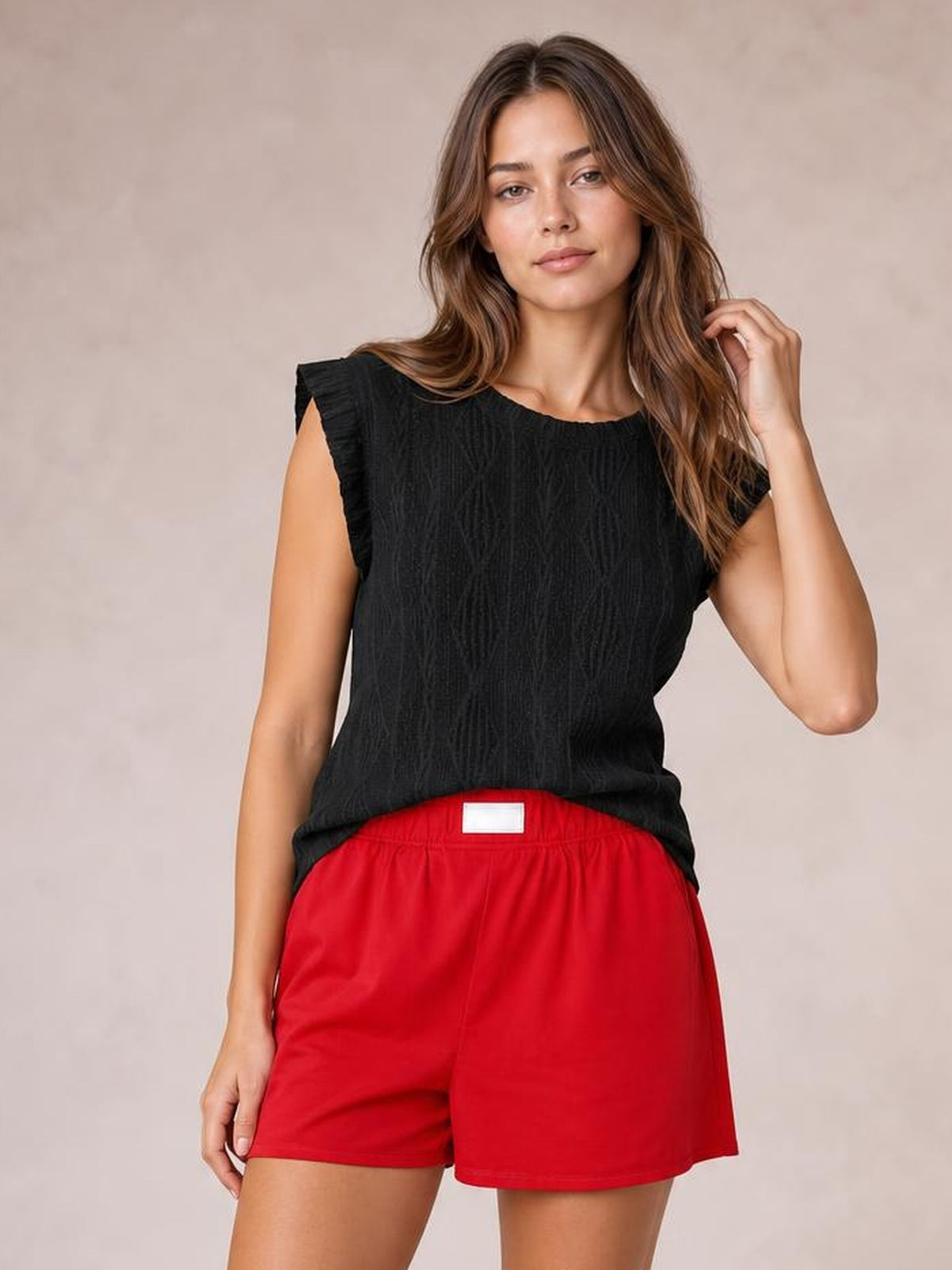 A woman modeling a black textured sleeveless top and bright red shorts against a neutral beige studio background.